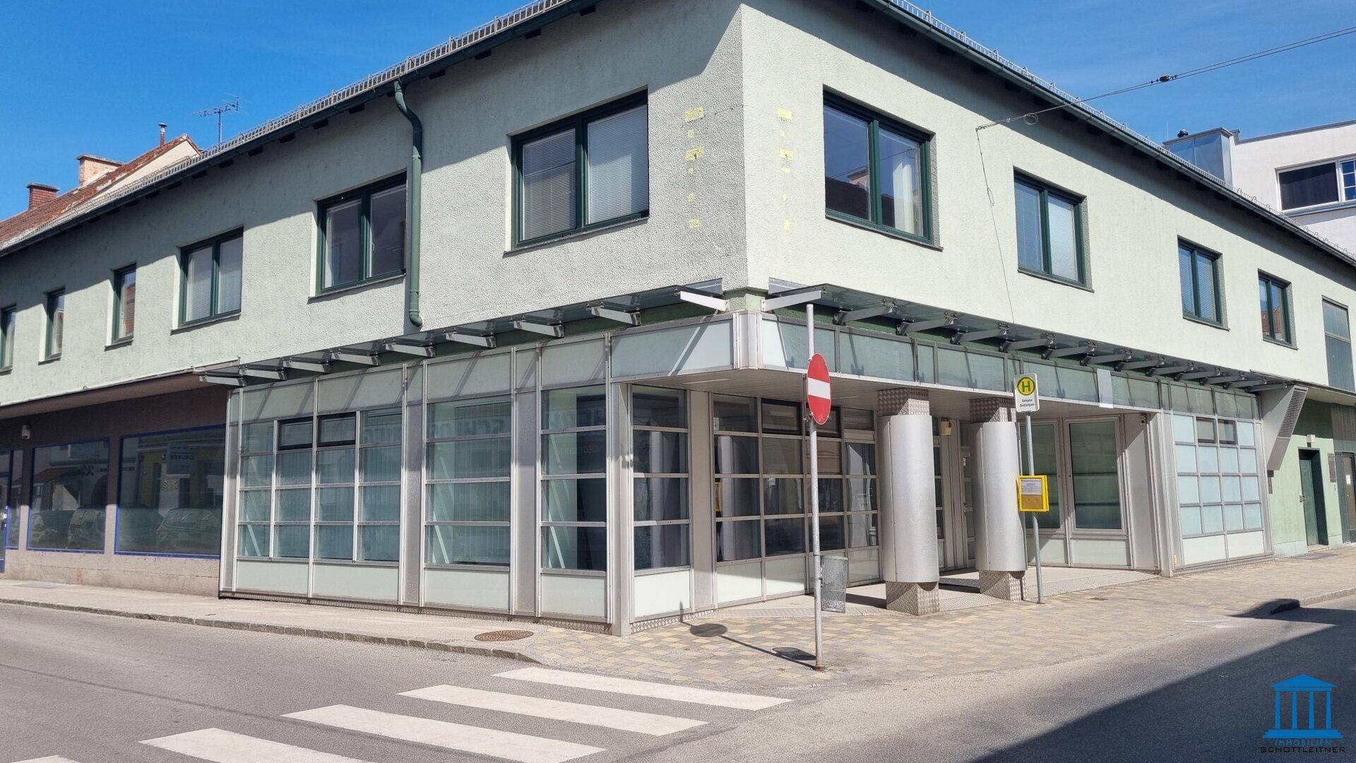 Weitwinkelansicht der Gebäudefassade an einer Straßenecke mit Zebrastreifen und Bushaltestelle.