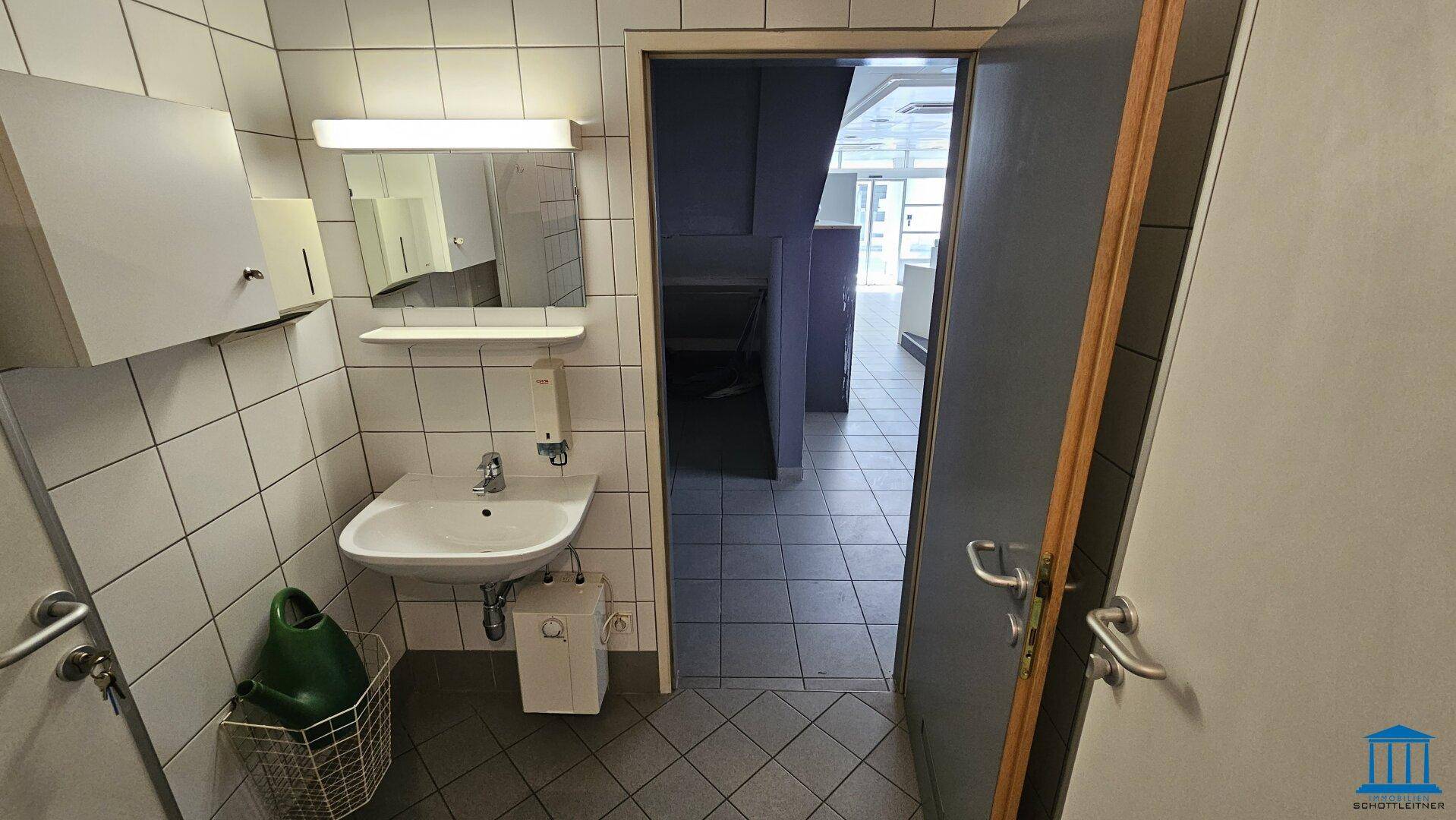 Waschbereich mit Waschbecken, Spiegel und weißen Fliesen, angrenzend an die Toiletten.