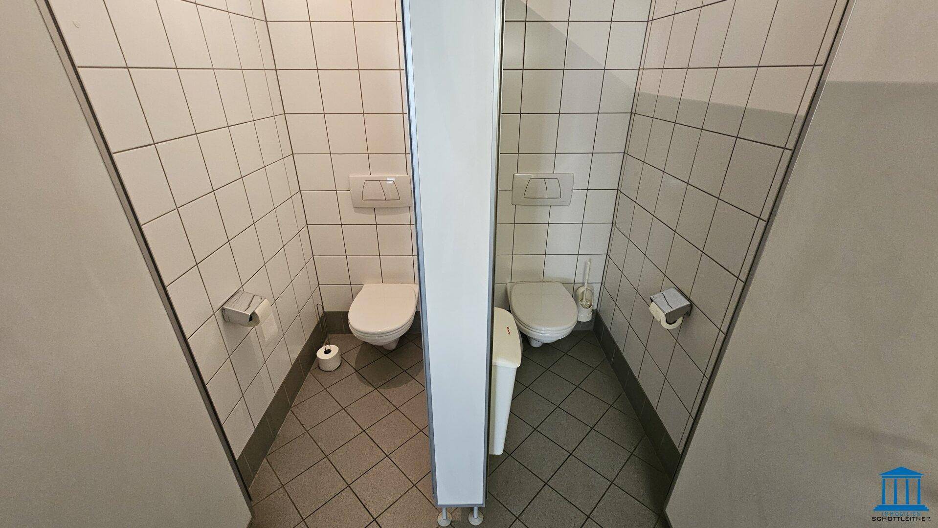 Zwei separate Toilettenkabinen mit weißen Fliesen und Wand-WCs in einem gepflegten Zustand.