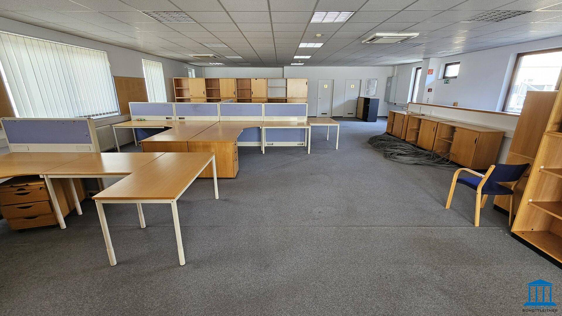 Weitläufiges Büro mit Teppichboden, Schreibtischen und Trennwänden, die Arbeitsbereiche definieren.