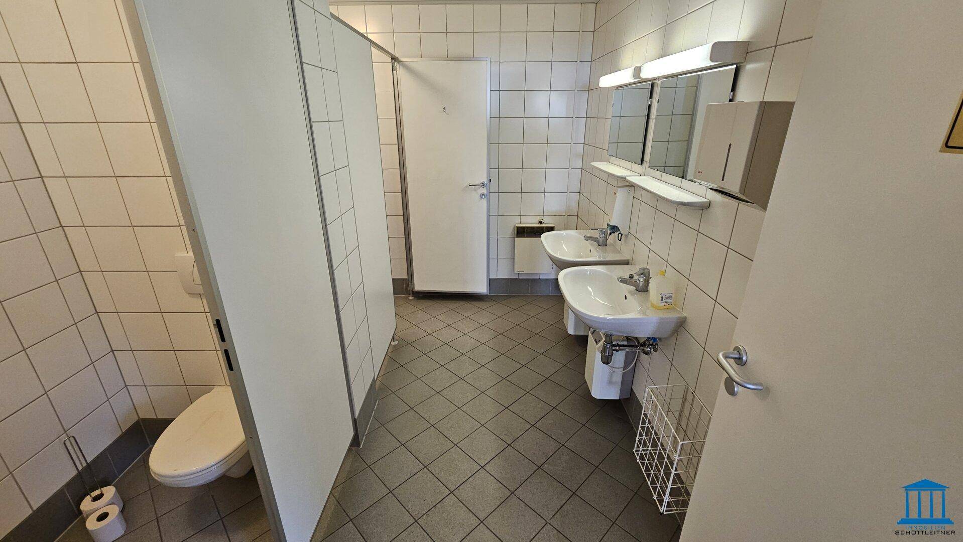 Öffentliche Toilettenanlage mit mehreren Kabinen, Waschbecken und gefliesten Wänden und Böden.