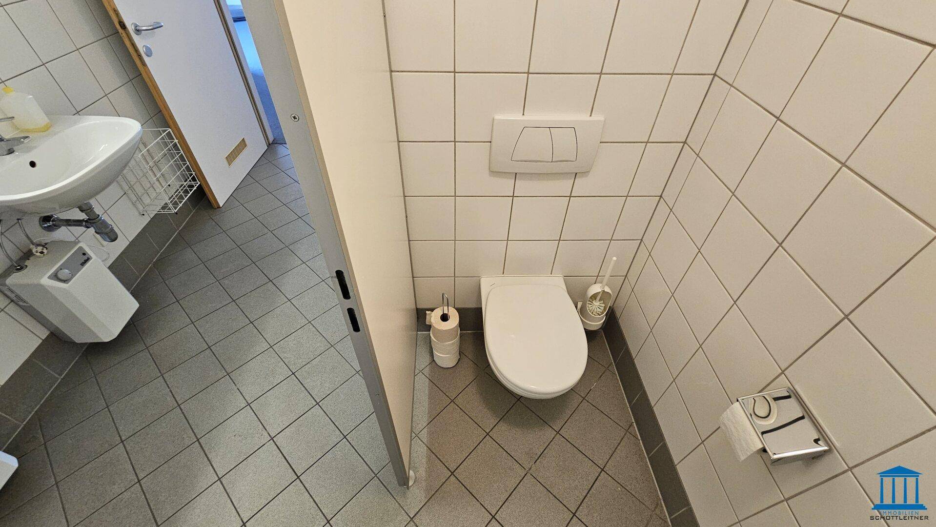 Einzelne Toilettenkabine mit Wand-WC, Spülkasten und gefliestem Boden und Wänden.