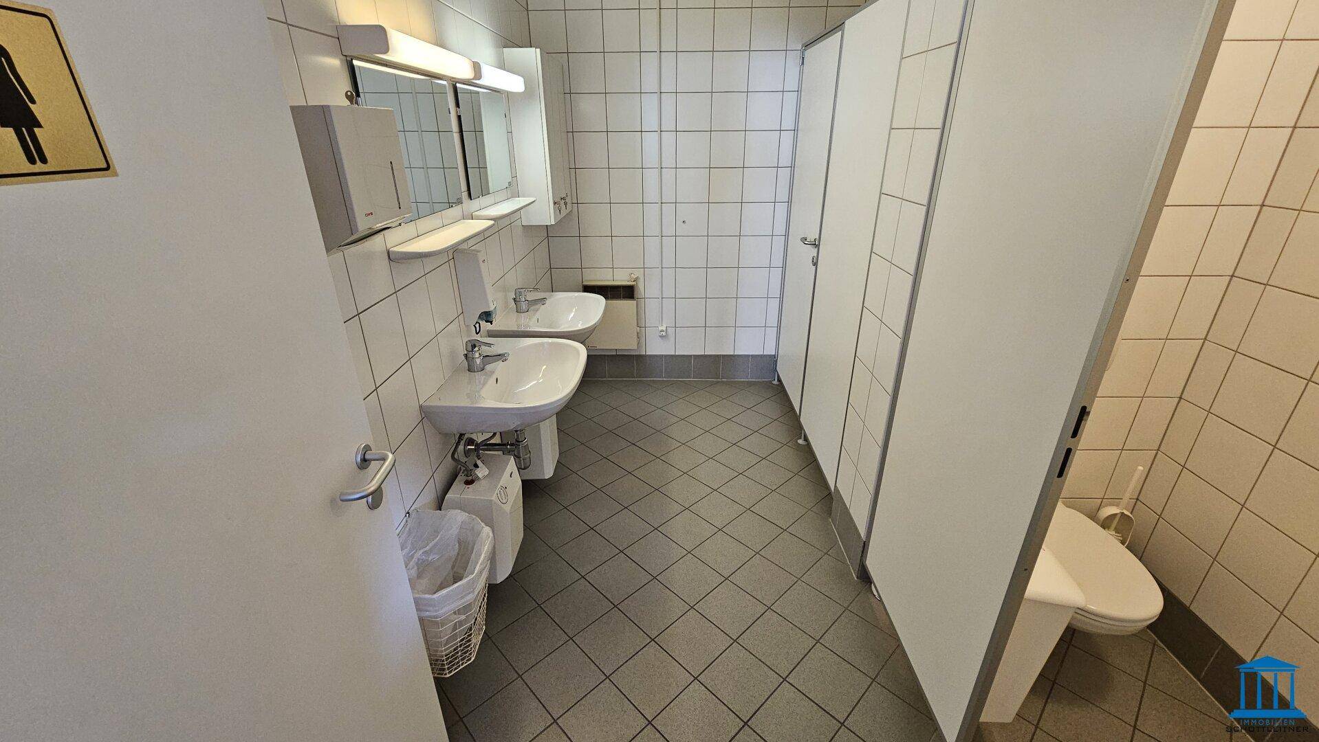 Sanitärbereich mit zwei Waschbecken, Spiegeln und Toilettenkabinen, alles gefliest und sauber.