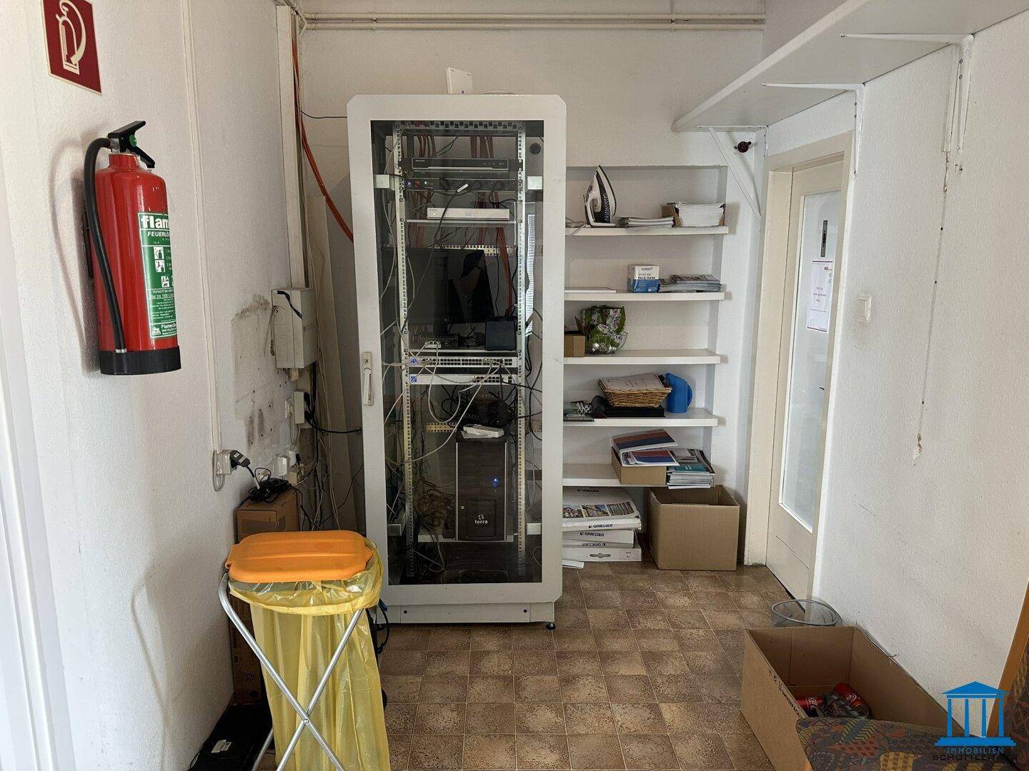Kleiner Raum mit einem Serverschrank, Regalen und einem Feuerlöscher an der Wand.