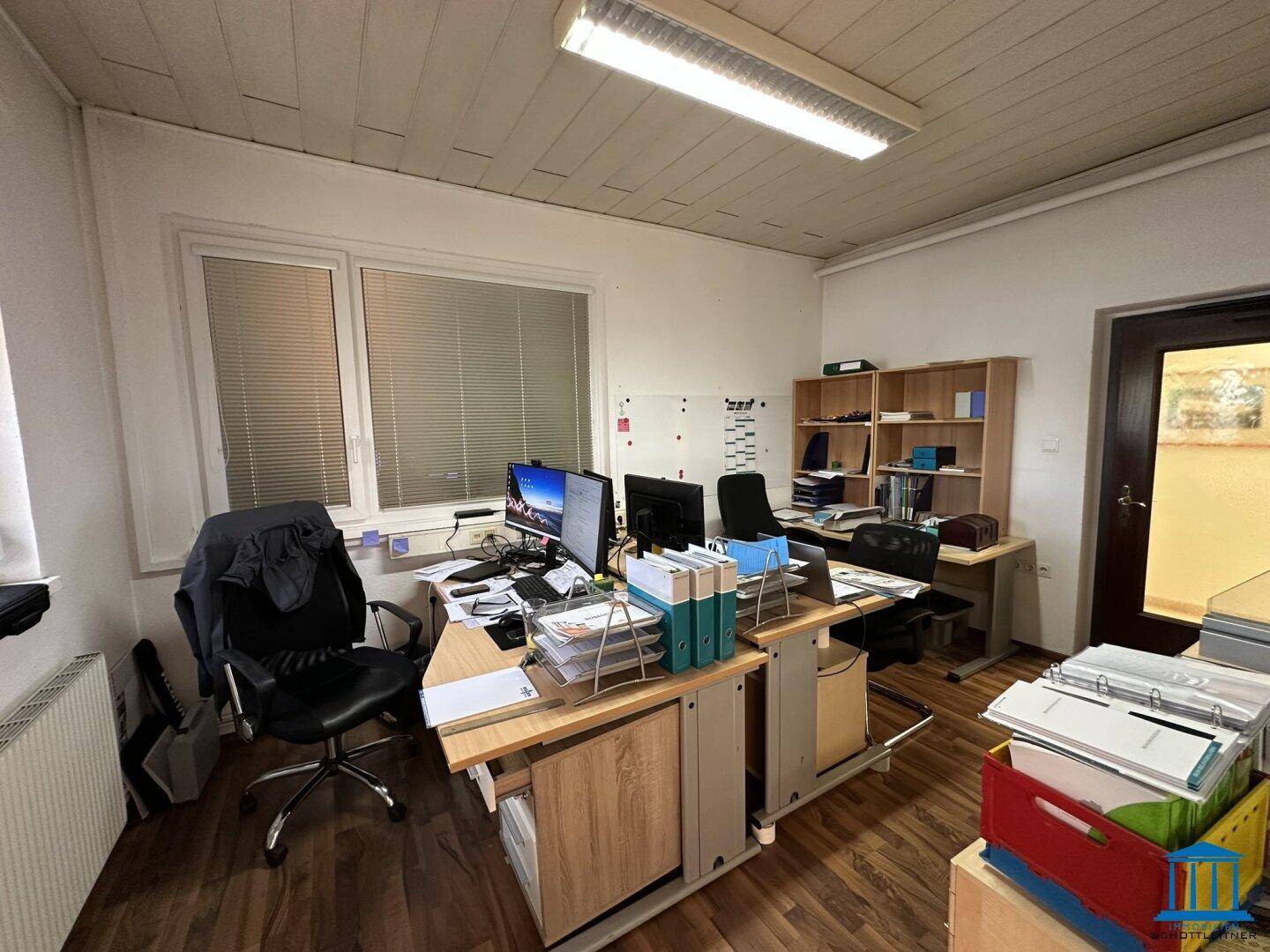Arbeitsplatz in einem Büro mit Schreibtisch, Computer und einem Fenster mit Jalousien.