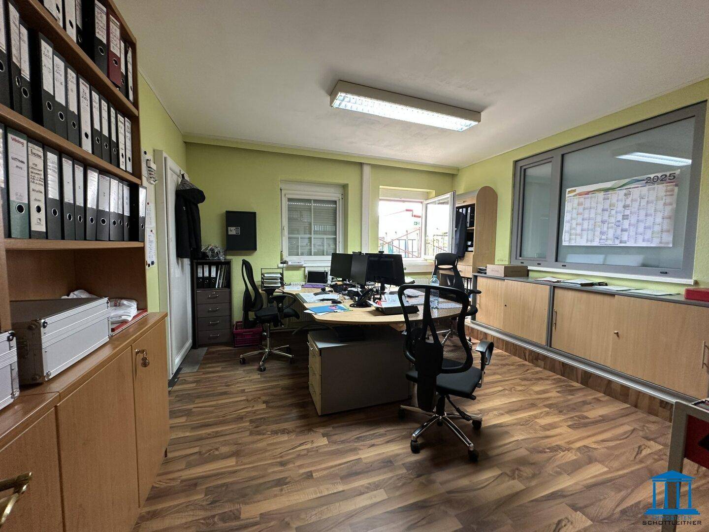 Büro mit vielen Aktenordnern, mehreren Bildschirmen und einem großen Fenster mit Blick nach draußen.