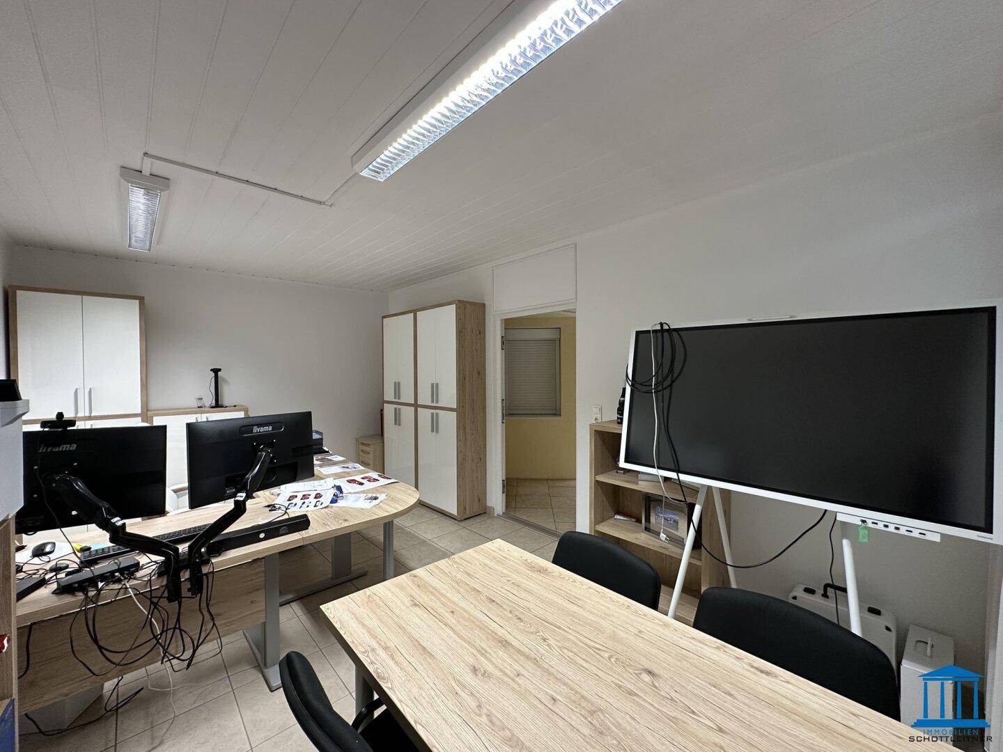 Geräumiges Büro mit mehreren Arbeitsplätzen, großem Bildschirm und hellen Schränken für Stauraum.