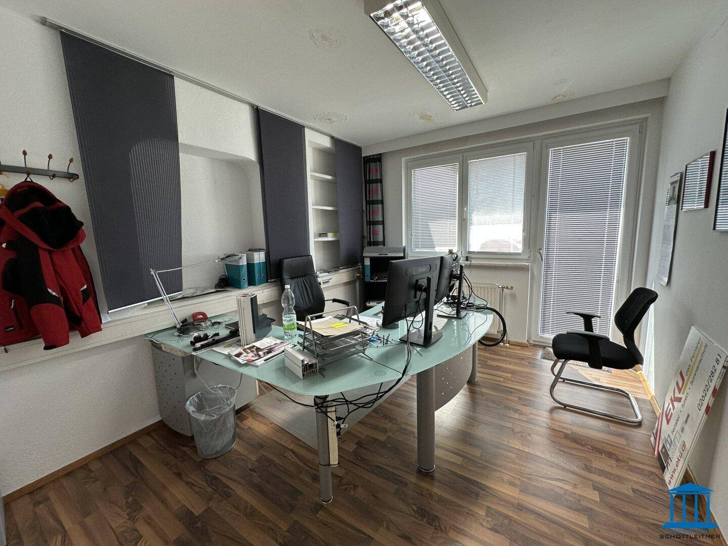 Helles Büro mit Schreibtisch, Bürostühlen und großen Fenstern mit Jalousien für angenehmes Arbeiten.