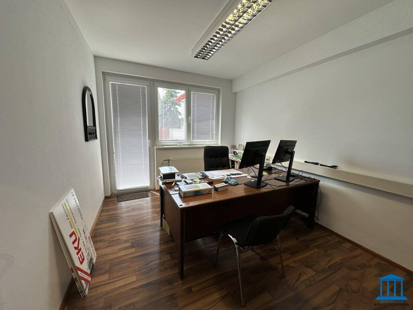 Einfaches Büro mit einem Schreibtisch, zwei Monitoren und einem Fenster mit Zugang nach draußen.