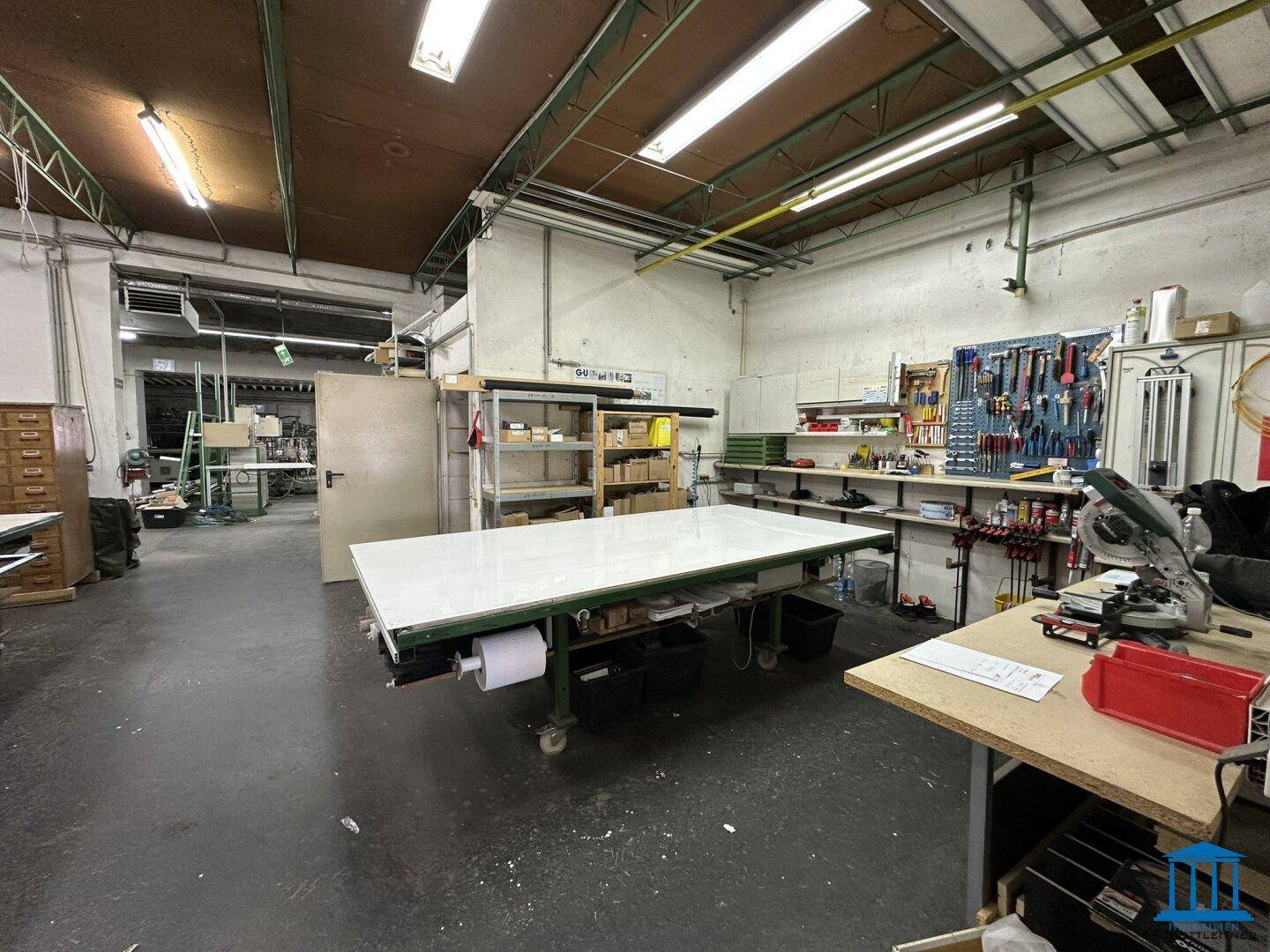 Werkstatt mit großen Arbeitstischen, Werkzeugen an der Wand und industrieller Beleuchtung.