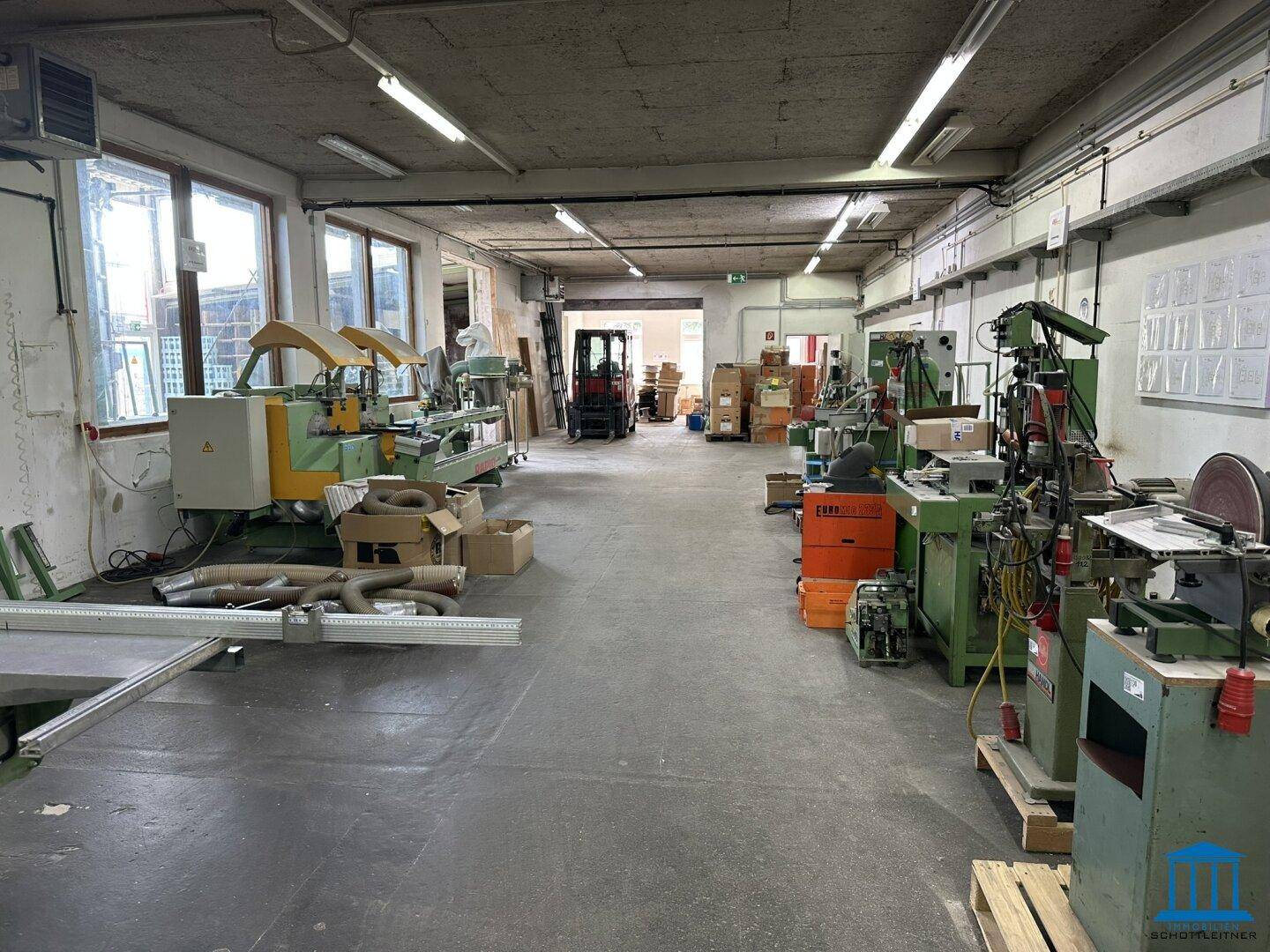Große Werkstatt mit verschiedenen Industriemaschinen, Fenstern und einem offenen Durchgang im Hintergrund.