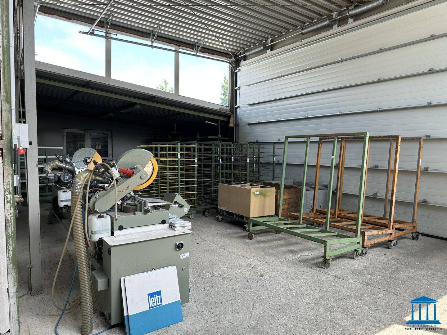 Werkstatt- und Lagerbereich mit einer großen Schneidemaschine, Metallregalen und einem Rolltor mit Fenstern.