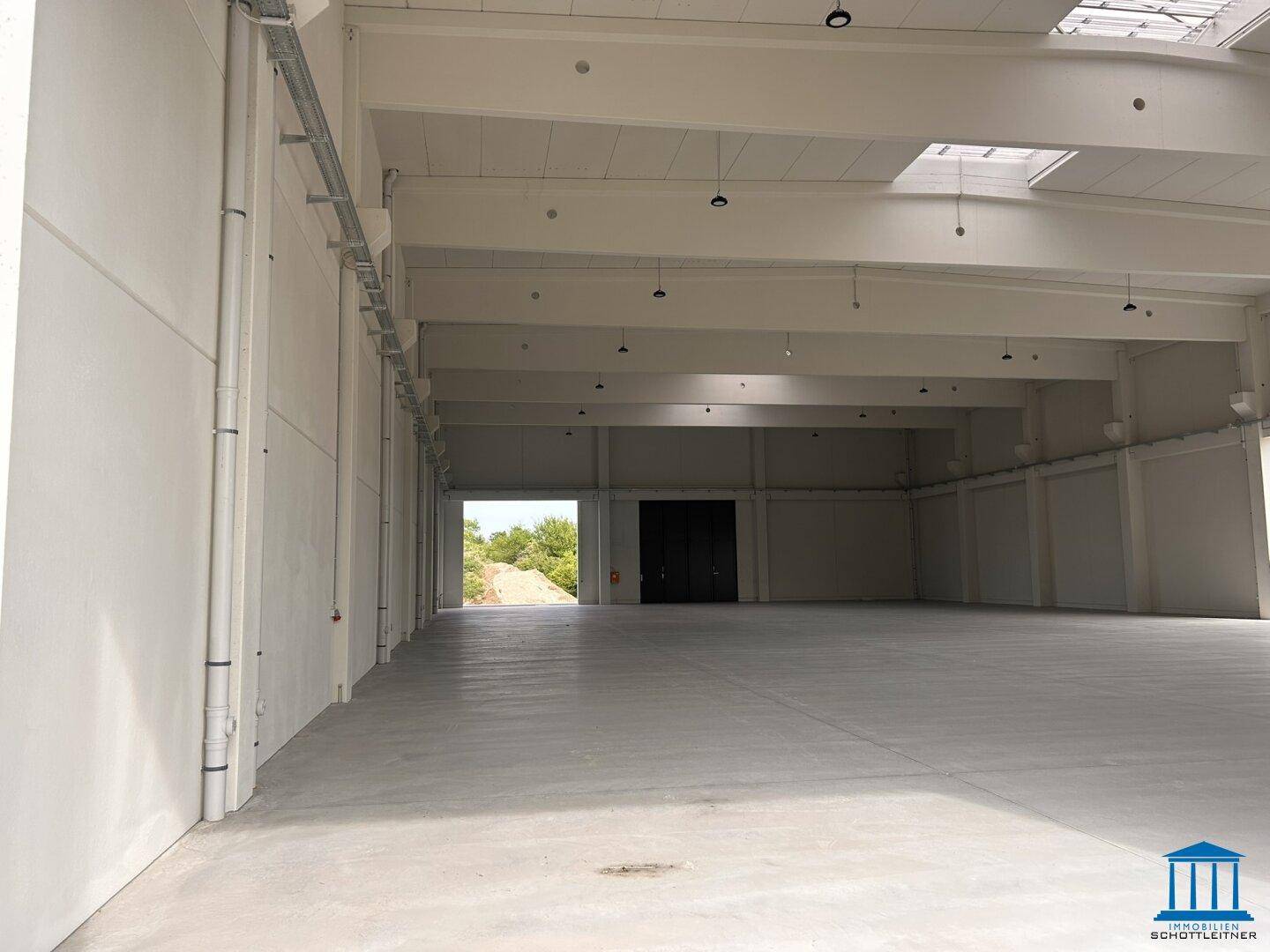 Leere Industriehalle mit hohen Wänden, Betonboden und Zugang zu einem Außenbereich.