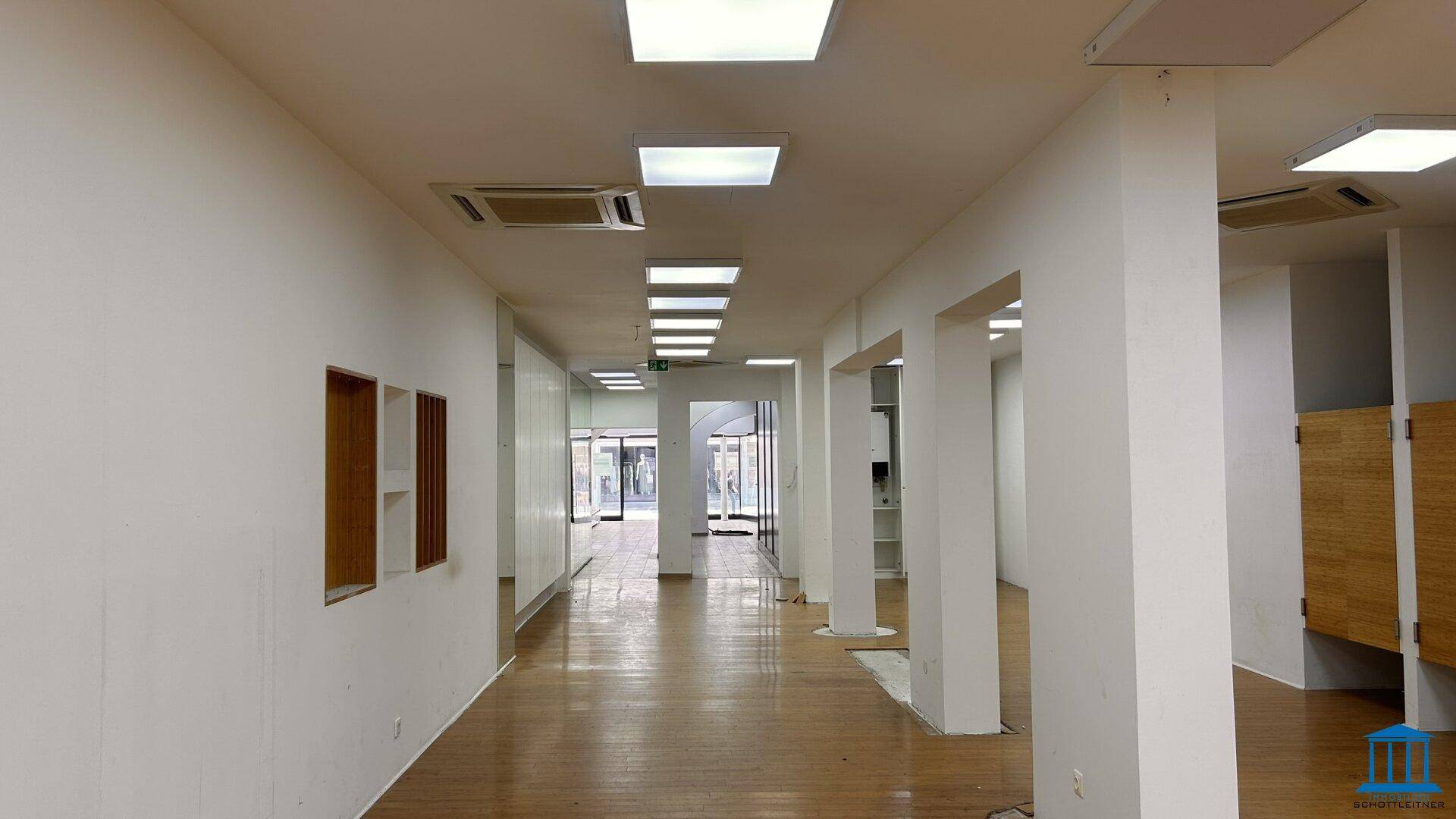 Geräumiger Verkaufsraum mit Holzfußboden, Säulen und zahlreichen Deckenleuchten für helle Ausleuchtung.
