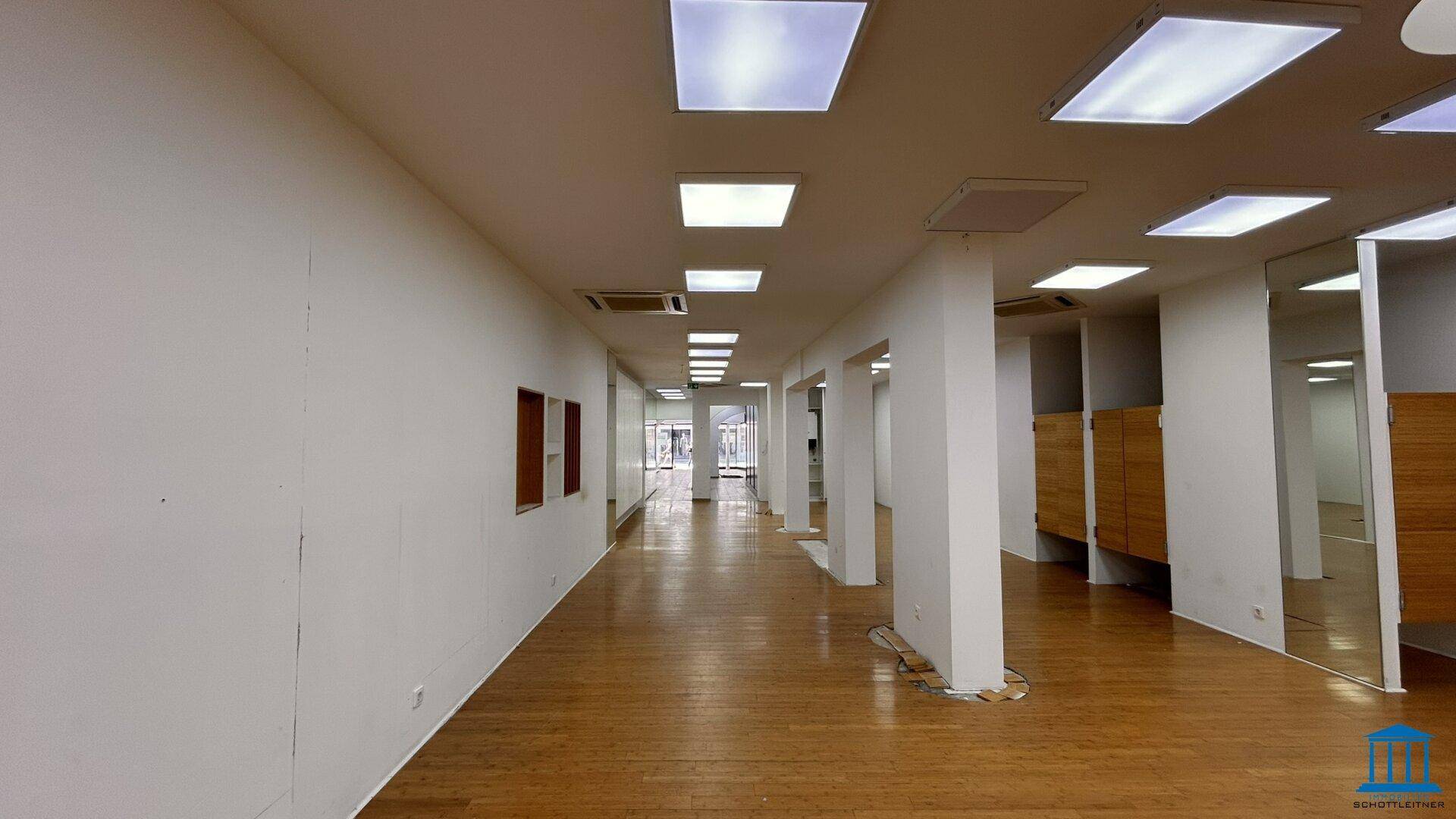 Langer, heller Verkaufsraum mit Holzfußboden, weißen Wänden und modernen Deckenleuchten.