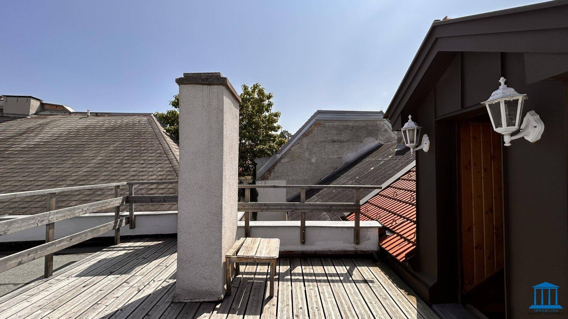 Dachterrasse mit Holzboden, Geländer und Blick auf umliegende Dächer und einen Schornstein unter klarem Himmel.