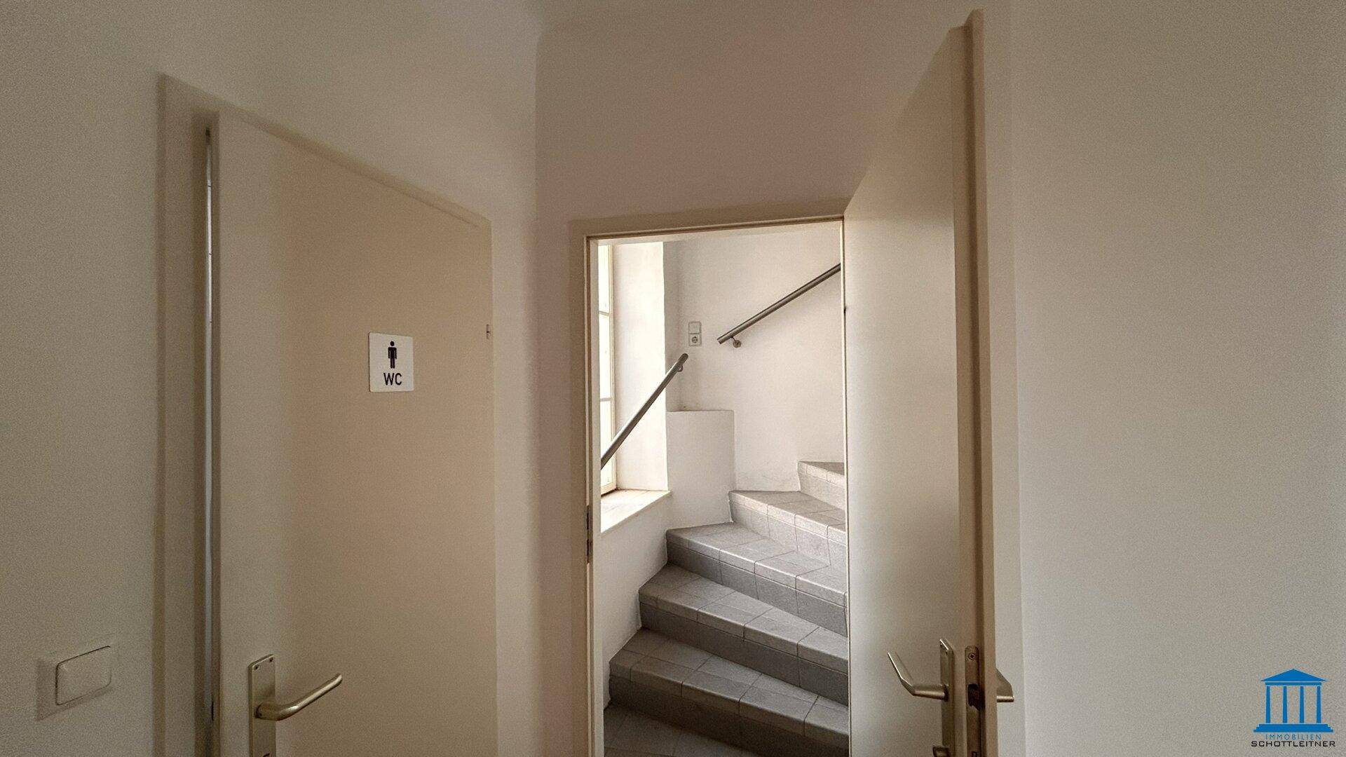 Heller Flur mit zwei Türen, eine davon zum WC, und Blick auf ein gefliestes Treppenhaus.