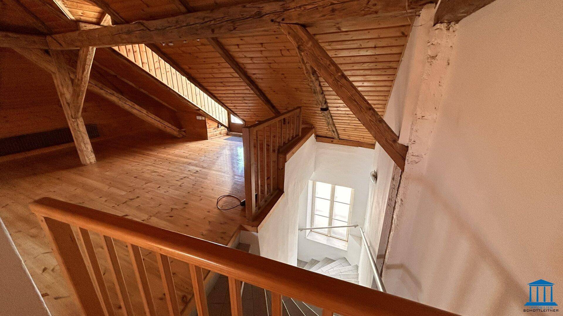 Dachbodenraum mit Holzboden und Treppenabgang, der zu einem hellen Treppenhaus mit Fenster führt.