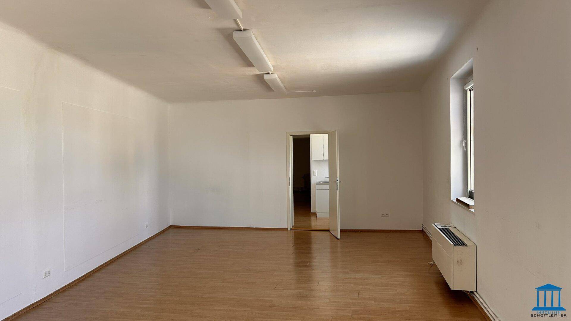 Leerer, heller Raum mit Holzboden, weißen Wänden und einem Fenster mit Heizkörper darunter.