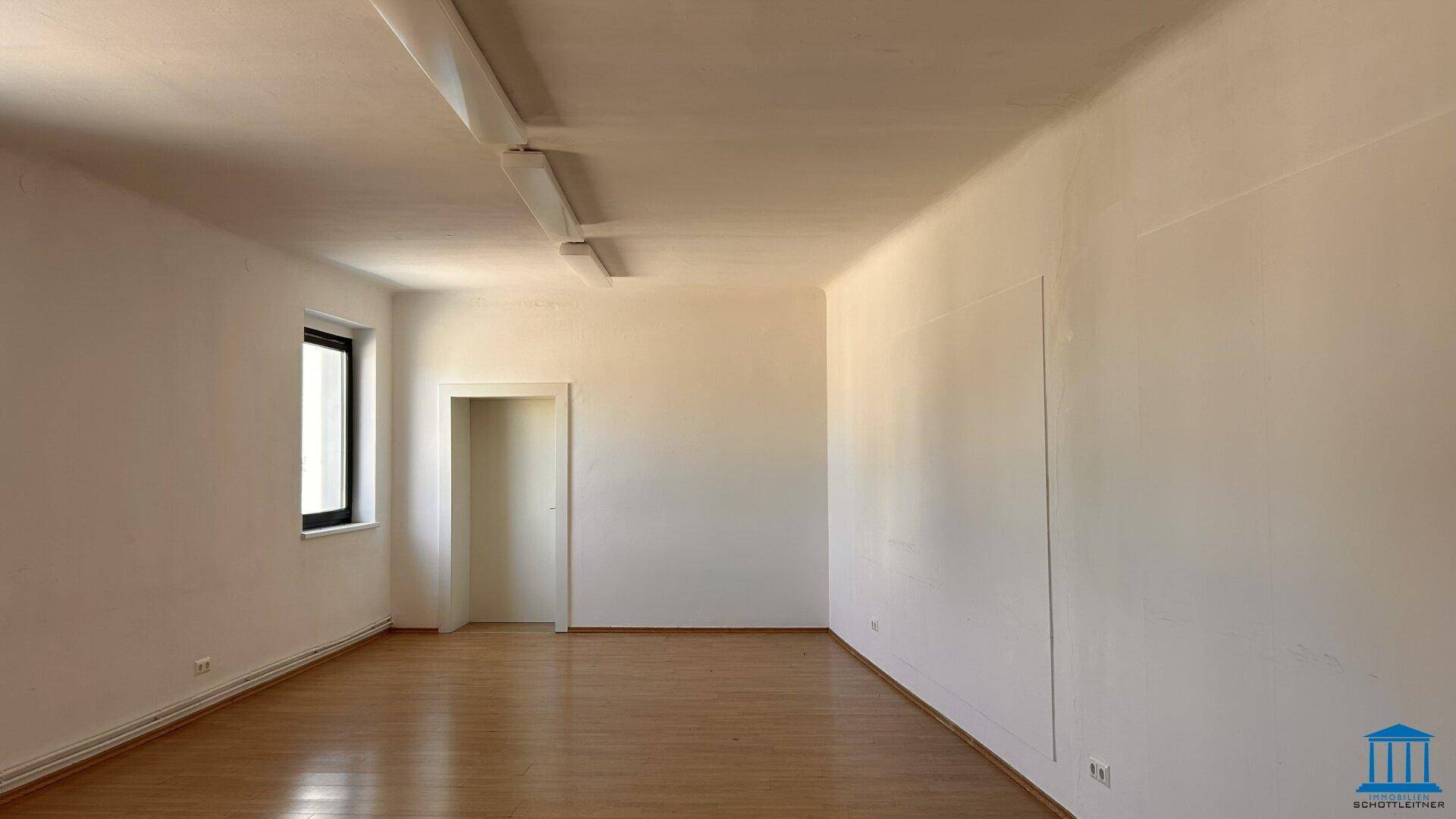 Heller, leerer Raum mit Holzboden, weißen Wänden und einem Fenster, das Tageslicht hereinlässt.