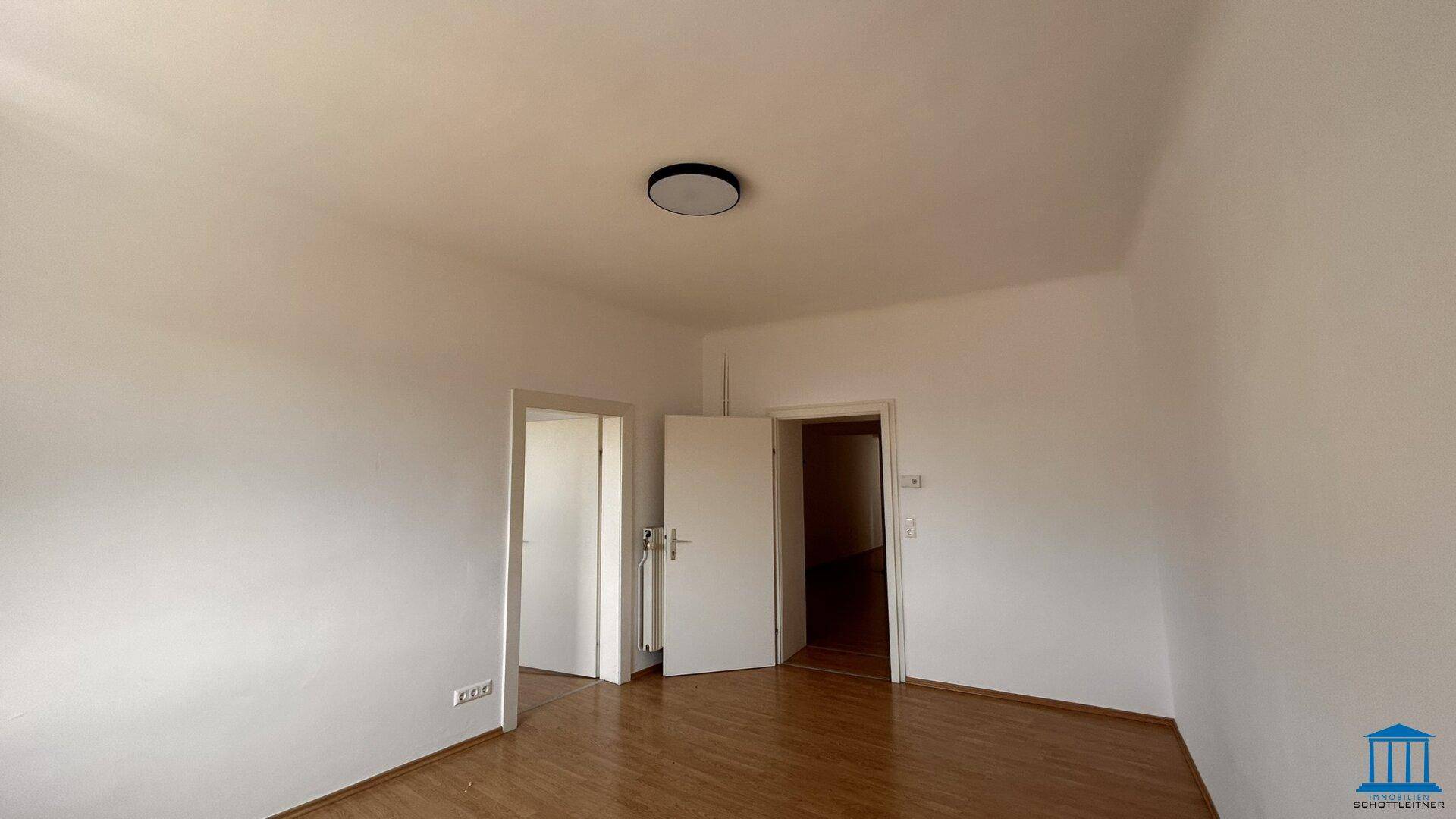 Heller, leerer Raum mit Holzboden, weißen Wänden und zwei Türen, die zu anderen Bereichen führen.