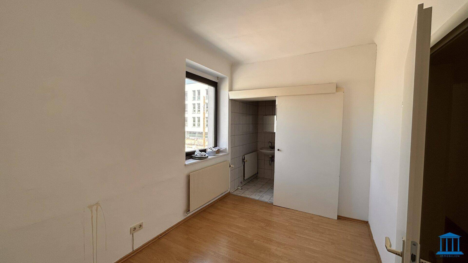 Heller Raum mit Holzboden, Fenster und einem abgetrennten Bereich, der eine kleine Küchenzeile beherbergen könnte.