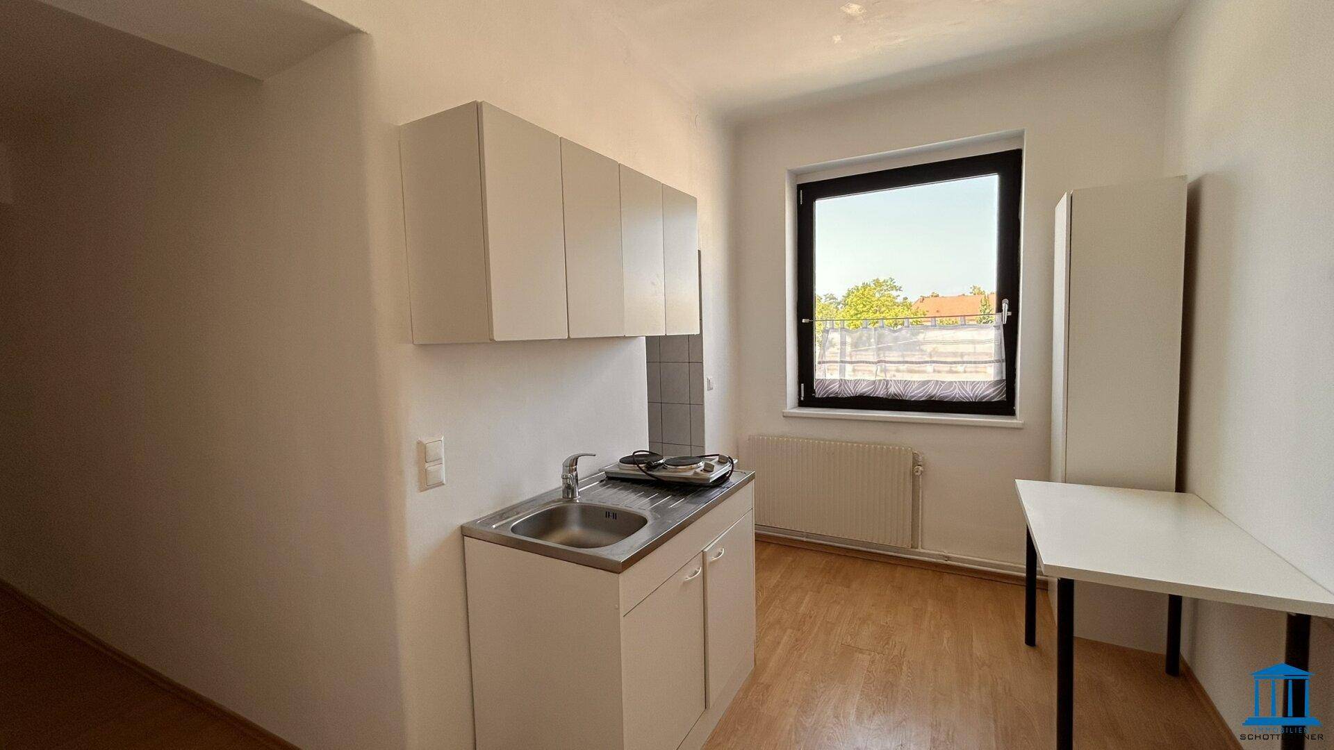Kleine Küchenzeile mit Spüle, Unterschränken und Hängeschränken, neben einem Fenster.