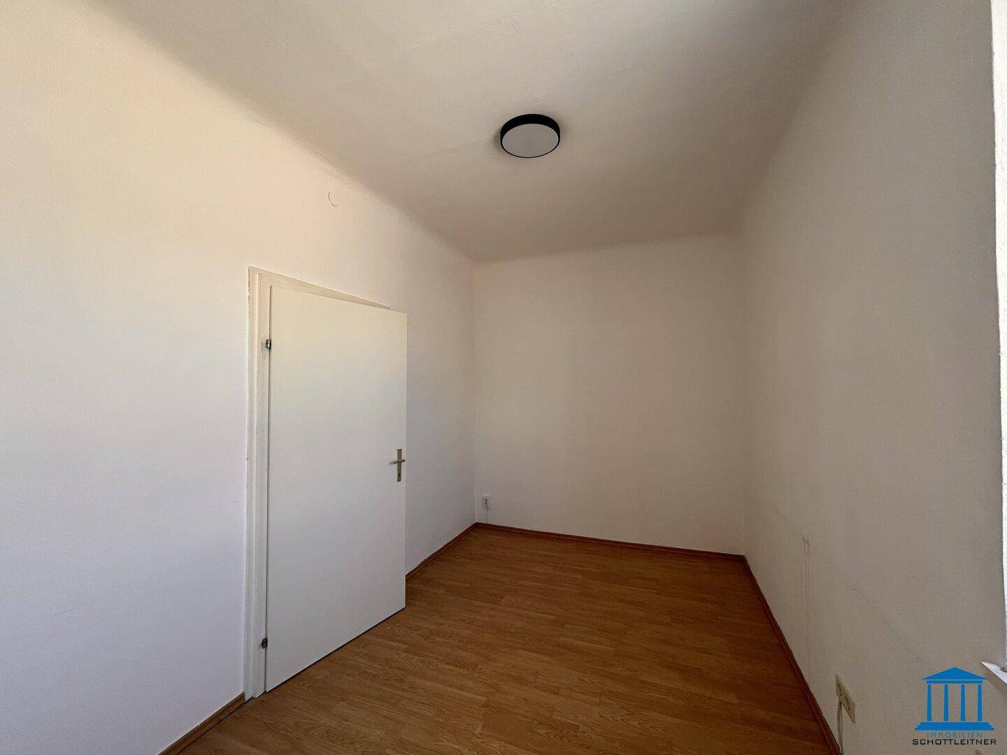 Kleiner, heller Raum mit Holzboden, weißen Wänden und einer Tür, ideal als Büro oder Abstellraum.