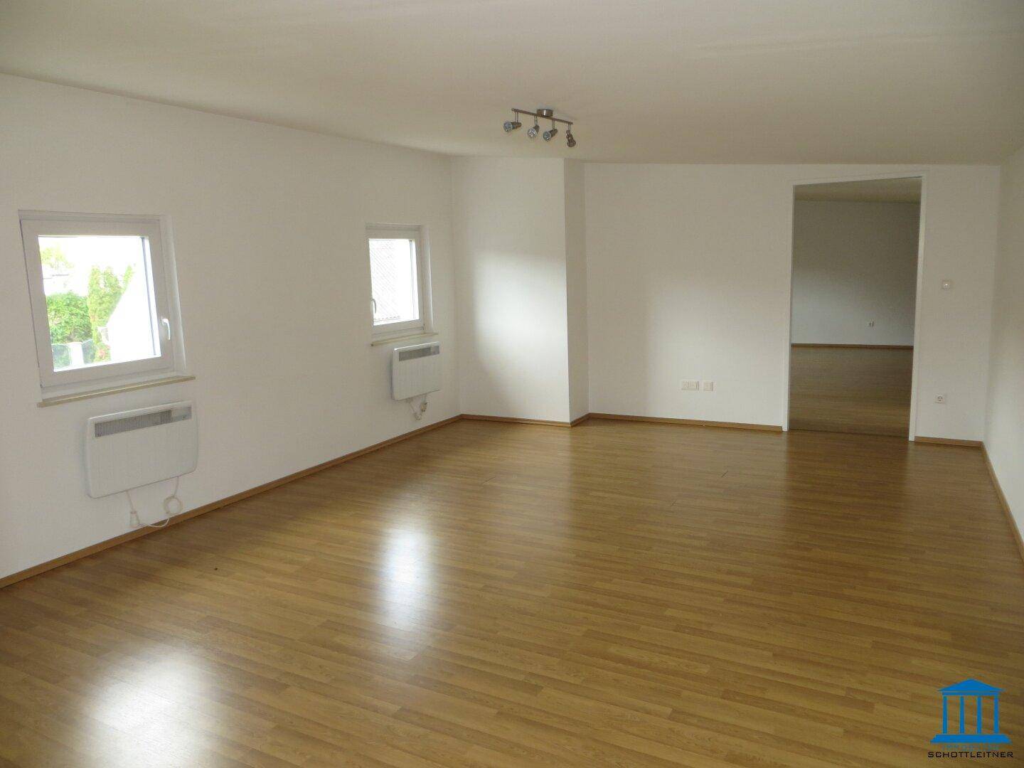 Geräumiges Wohnzimmer mit hellem Parkettboden, weißen Wänden und zwei Fenstern.