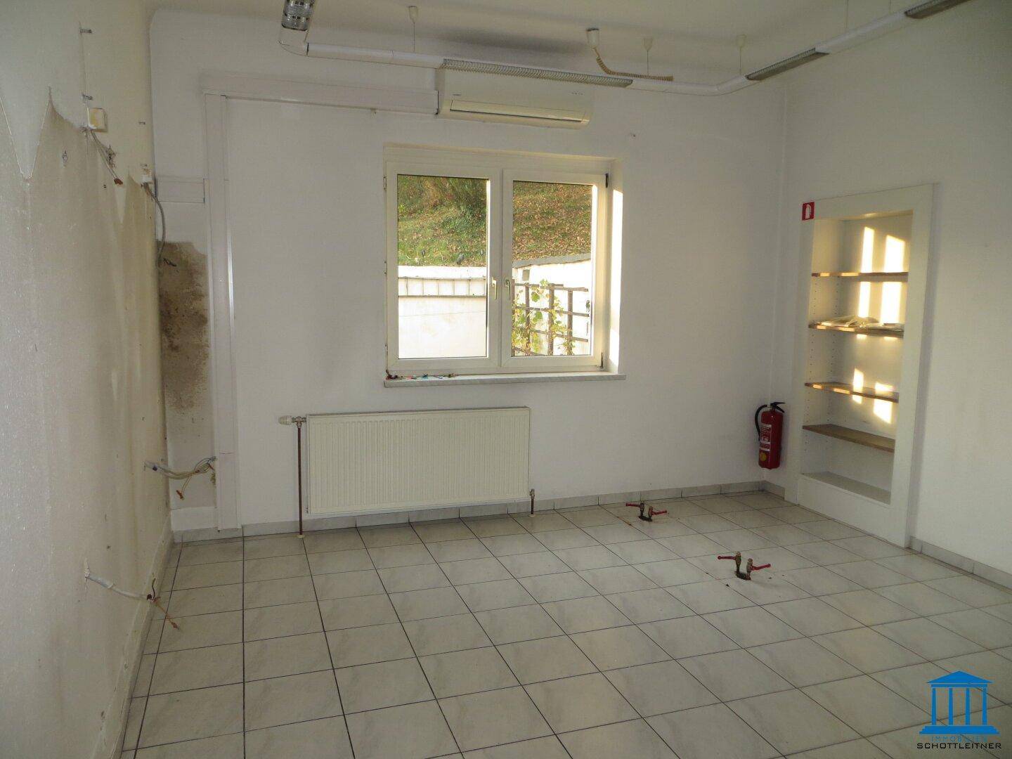 Heller Gewerberaum mit Fenster, Heizkörper und eingebauten Regalen, der Boden ist gefliest und die Wände sind renovierungsbedürftig.