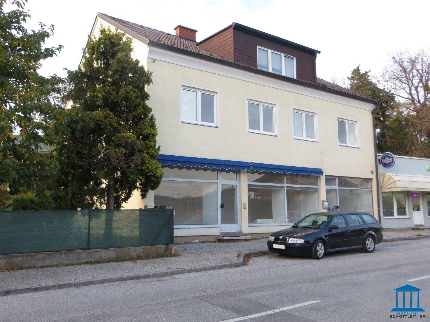 Breitere Außenansicht des Gebäudes mit Ladenlokalen im Erdgeschoss, umgeben von Bäumen und einer Straße mit parkendem Auto.
