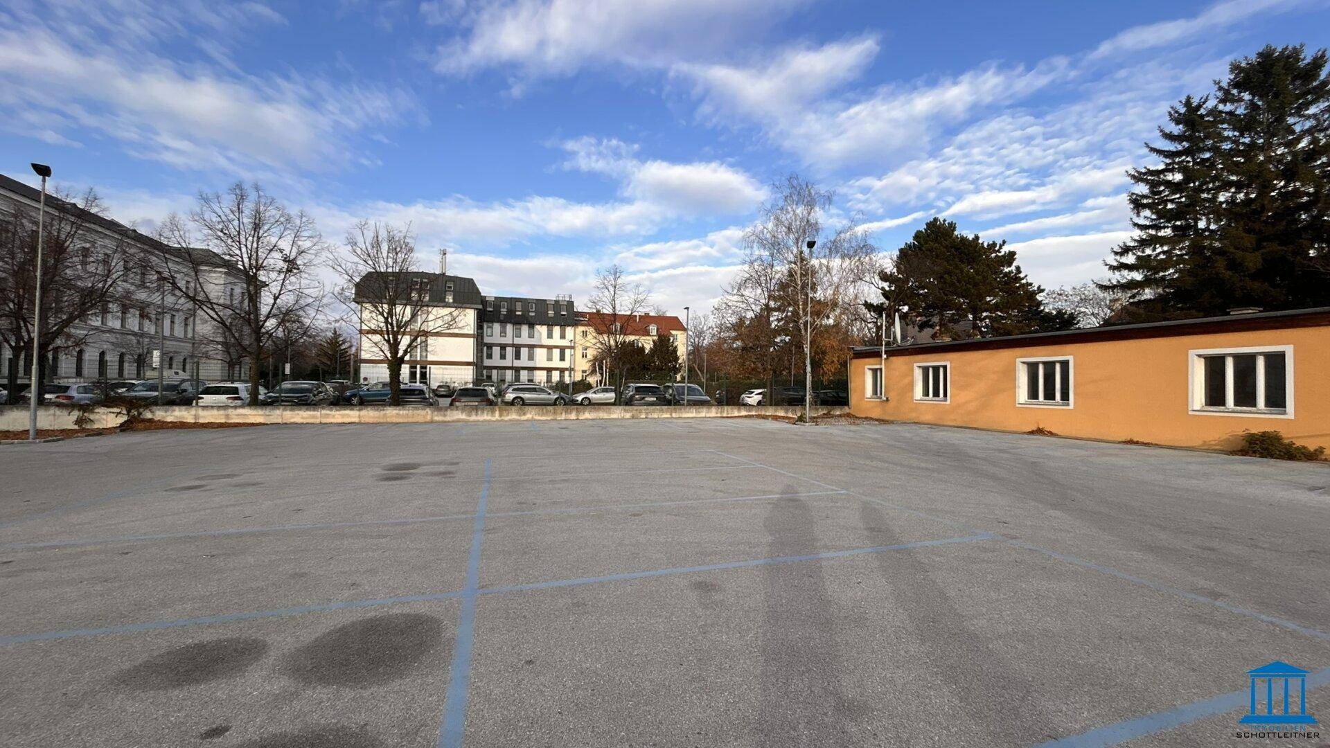 Großer Parkplatz mit blauen Markierungen und Gebäuden im Hintergrund unter bewölktem Himmel.