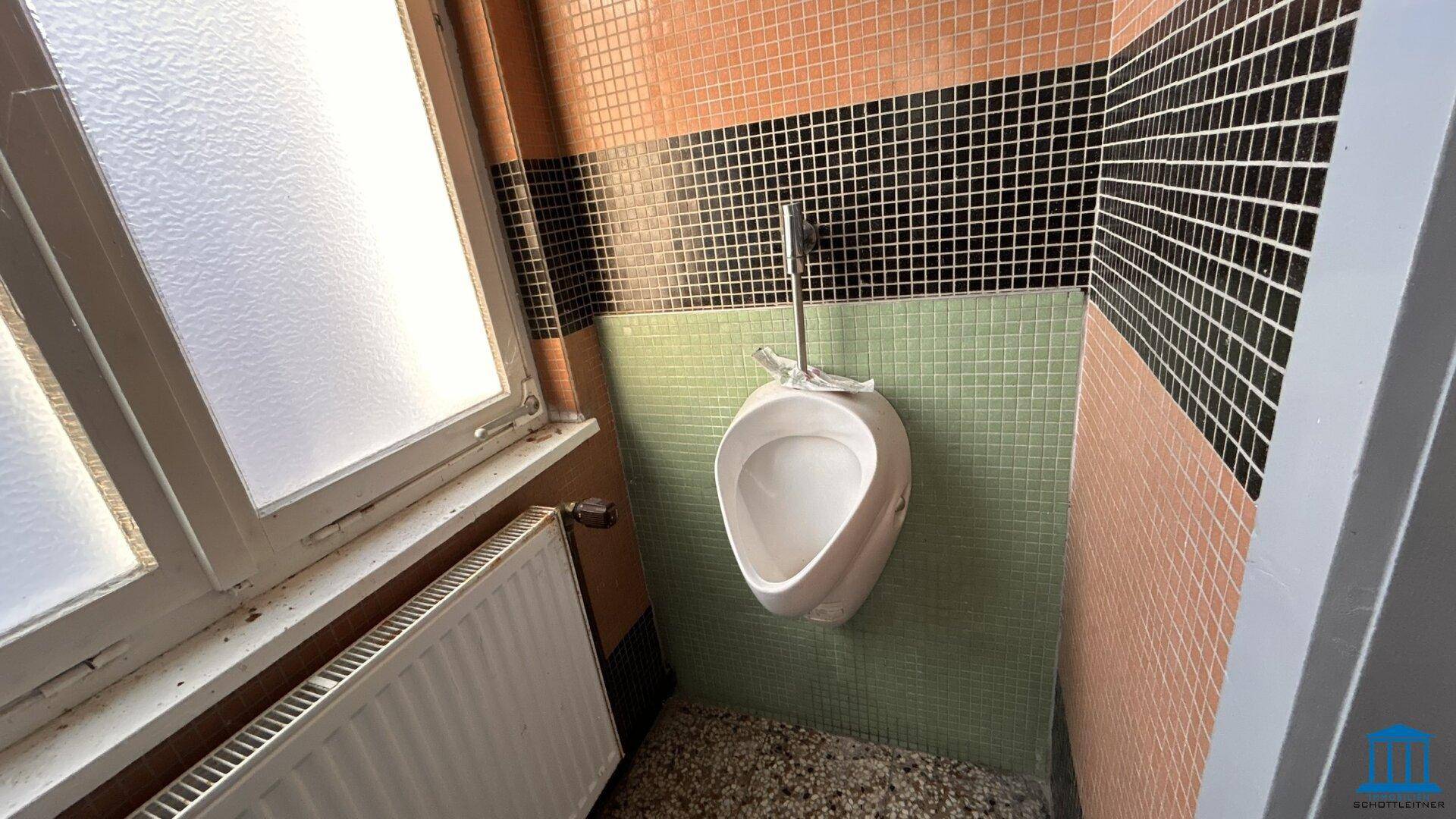 Badezimmer mit Urinal, farbigen Fliesen und einem Fenster, das natürliches Licht hereinlässt.