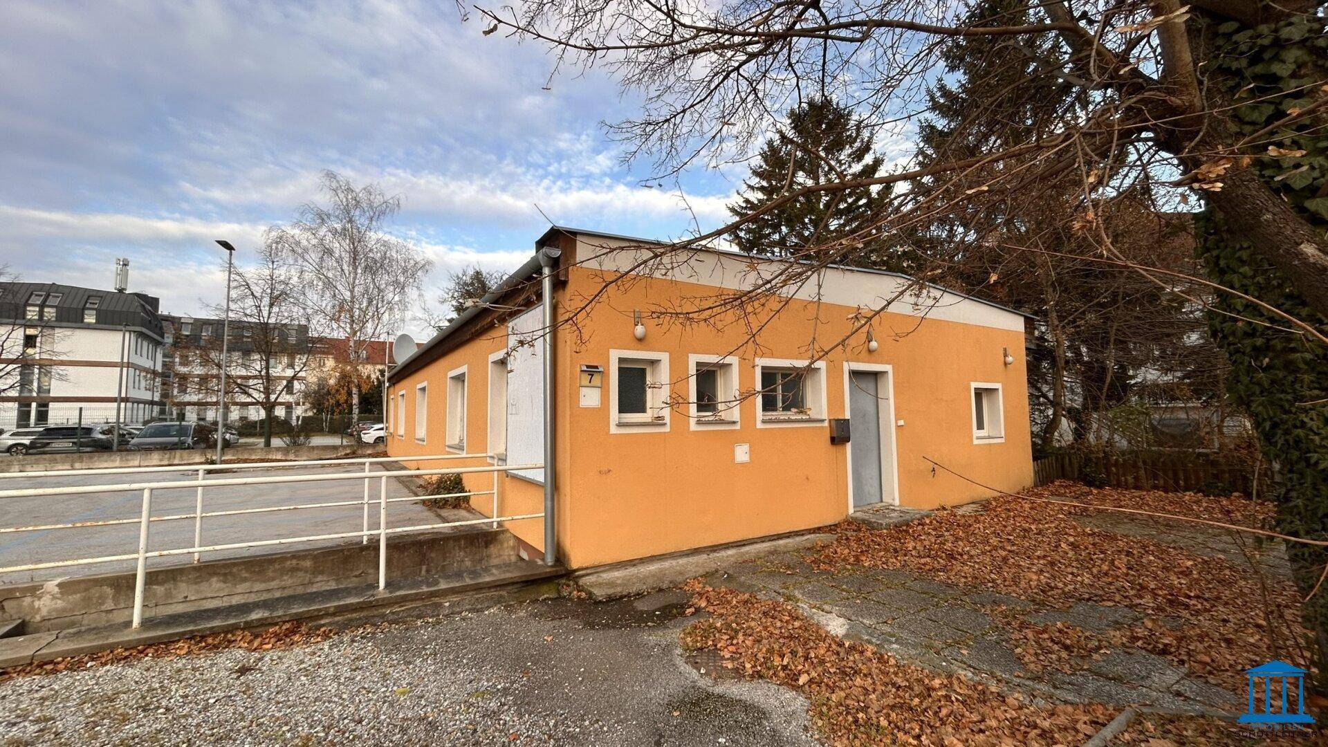 Seitenansicht eines gelben Gebäudes mit Eingangsbereich, Rampe und umgebender Vegetation.