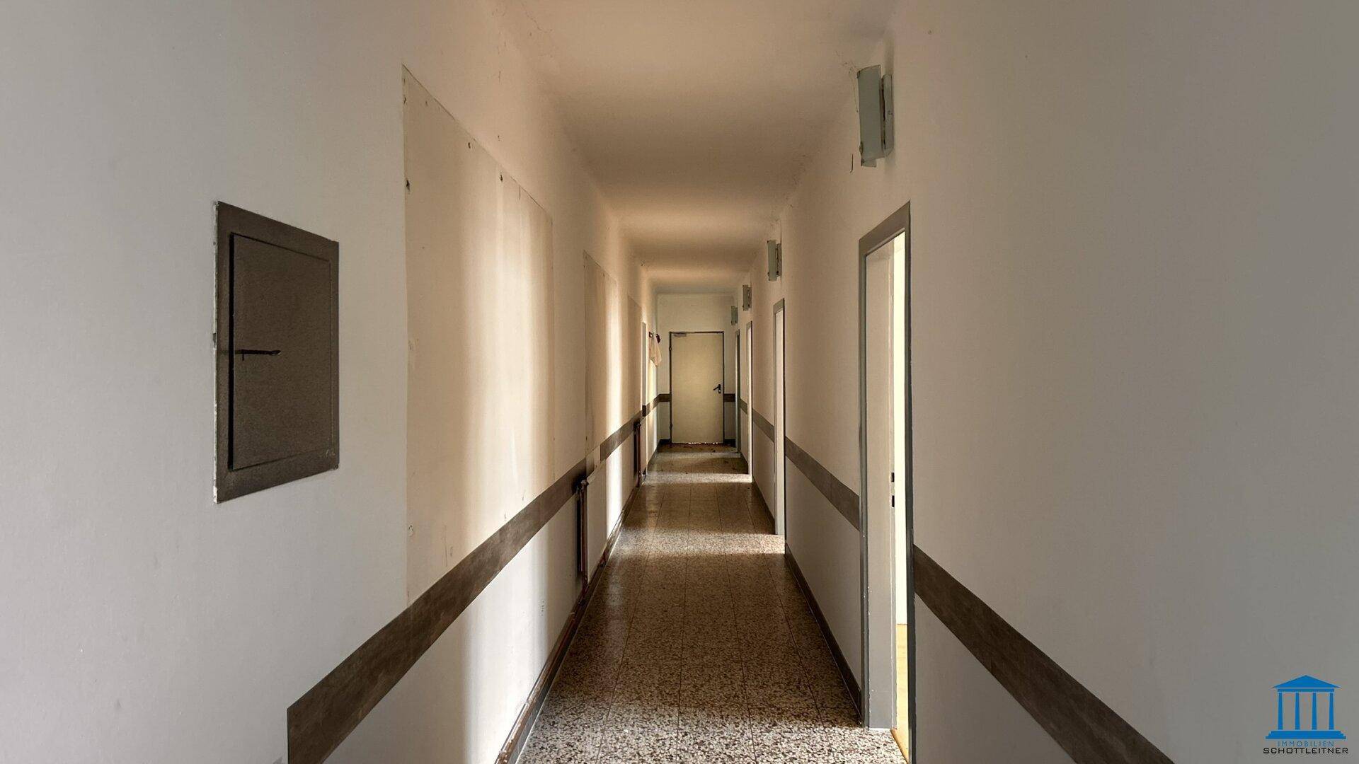 Langer, leerer Flur mit Terrazzoboden, weißen Wänden und mehreren Türen.