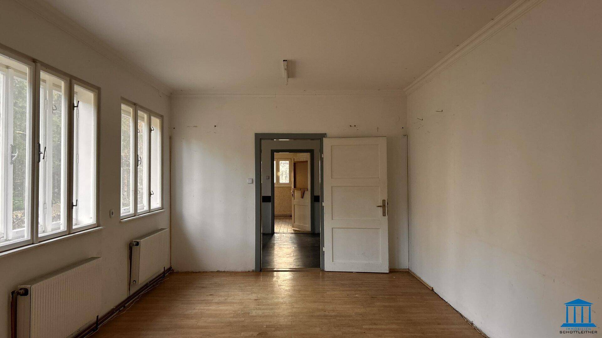 Geräumiger, leerer Raum mit Holzfußboden und mehreren Fenstern, die viel Licht hereinlassen.