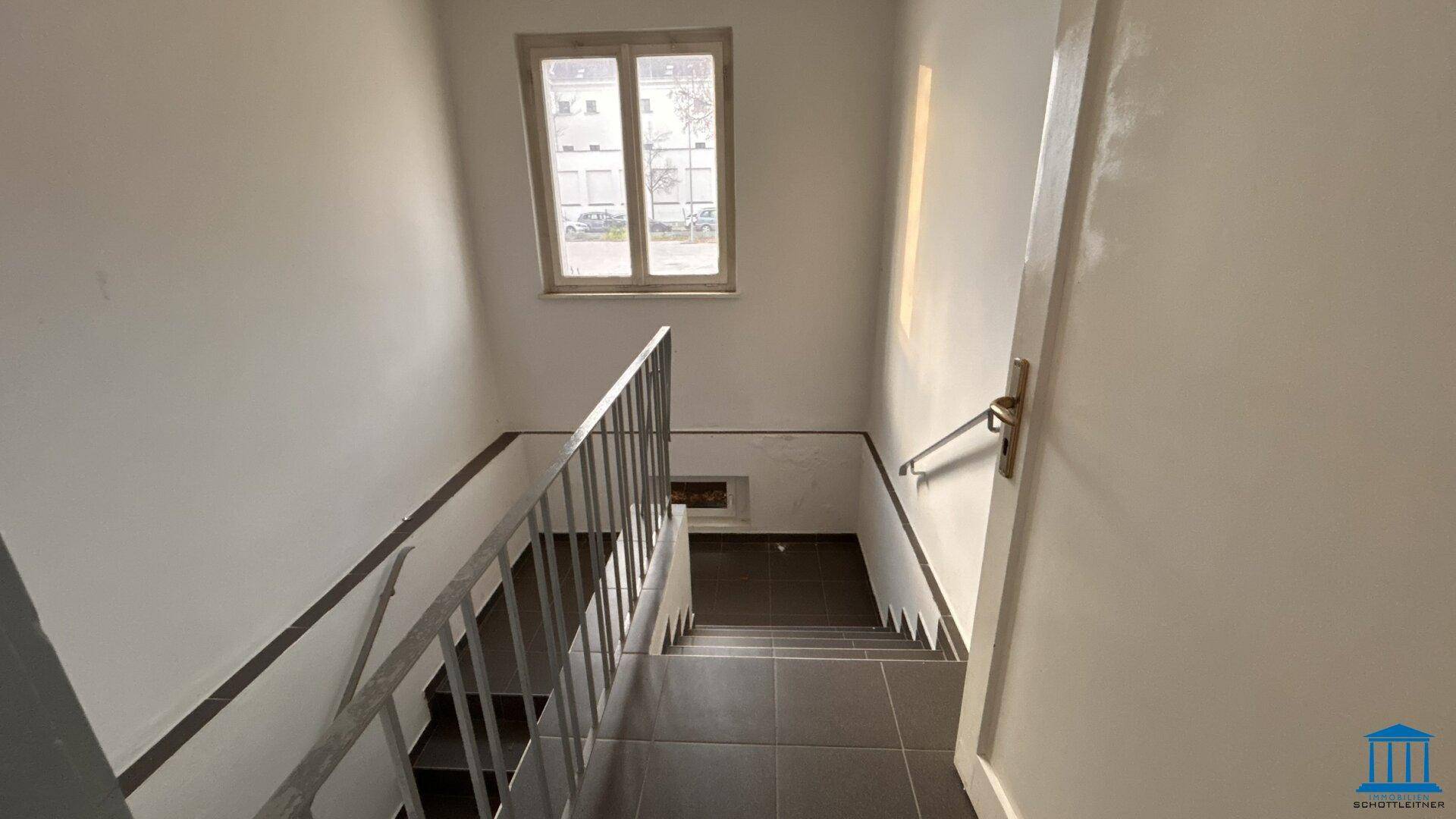 Treppenhaus mit Metallgeländer, gefliestem Boden und einem Fenster, das den Bereich erhellt.