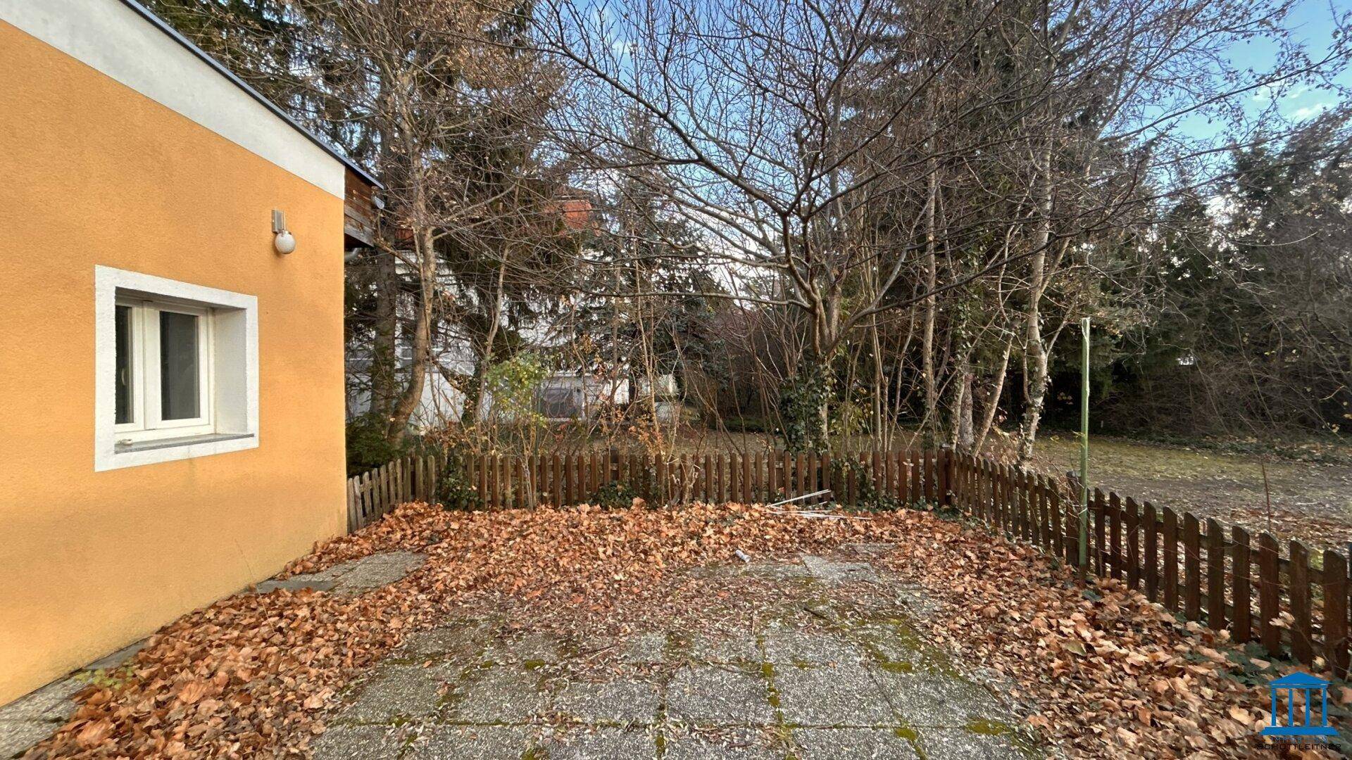 Gepflasterter Bereich neben dem Gebäude mit einem Holzzaun und Blick auf den Garten.