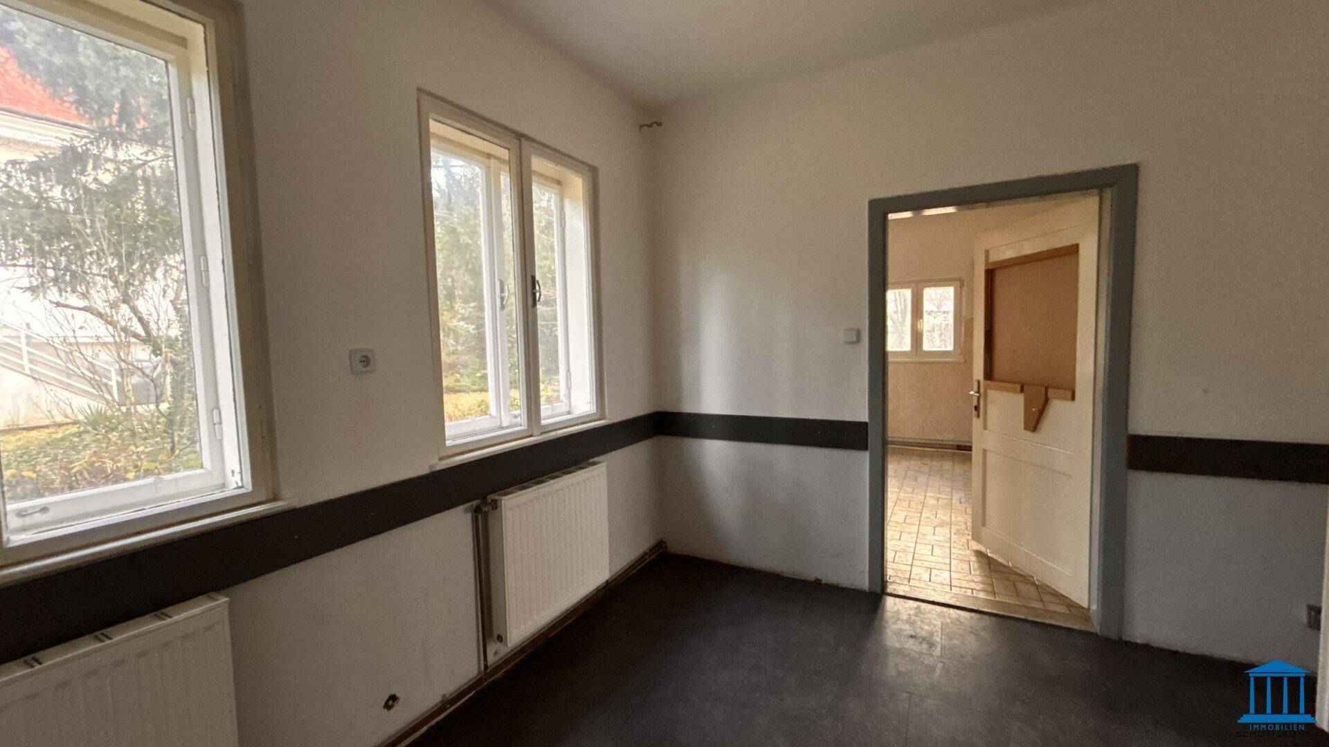 Leerer Raum mit zwei Fenstern, Heizkörpern und dunklem Boden, der Renovierung benötigt.