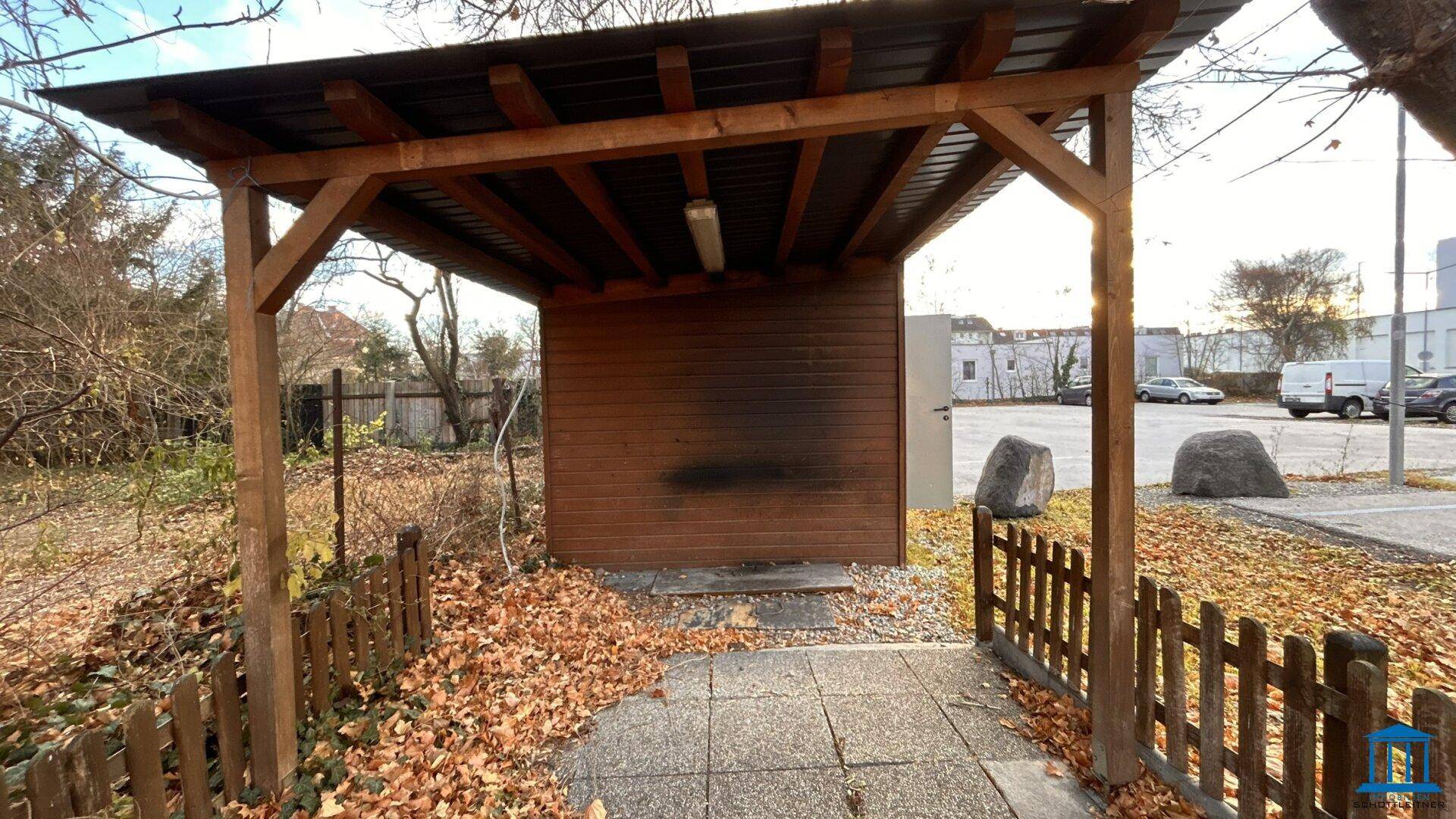 Überdachter Holzunterstand mit Blick auf einen Parkplatz und weitere Gebäude im Hintergrund.