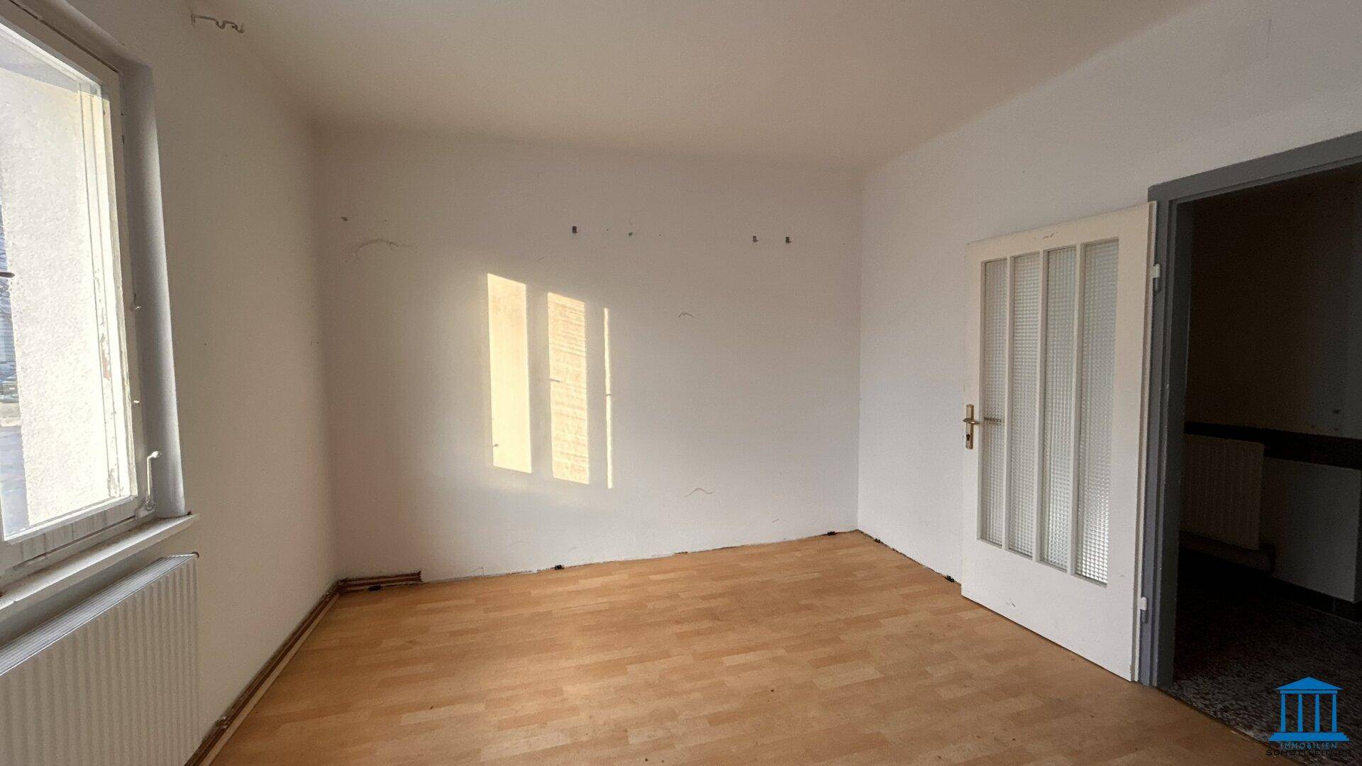 Leerer Raum mit Holzfußboden, weiß gestrichenen Wänden und einem Fenster, das Sonnenlicht hereinlässt.