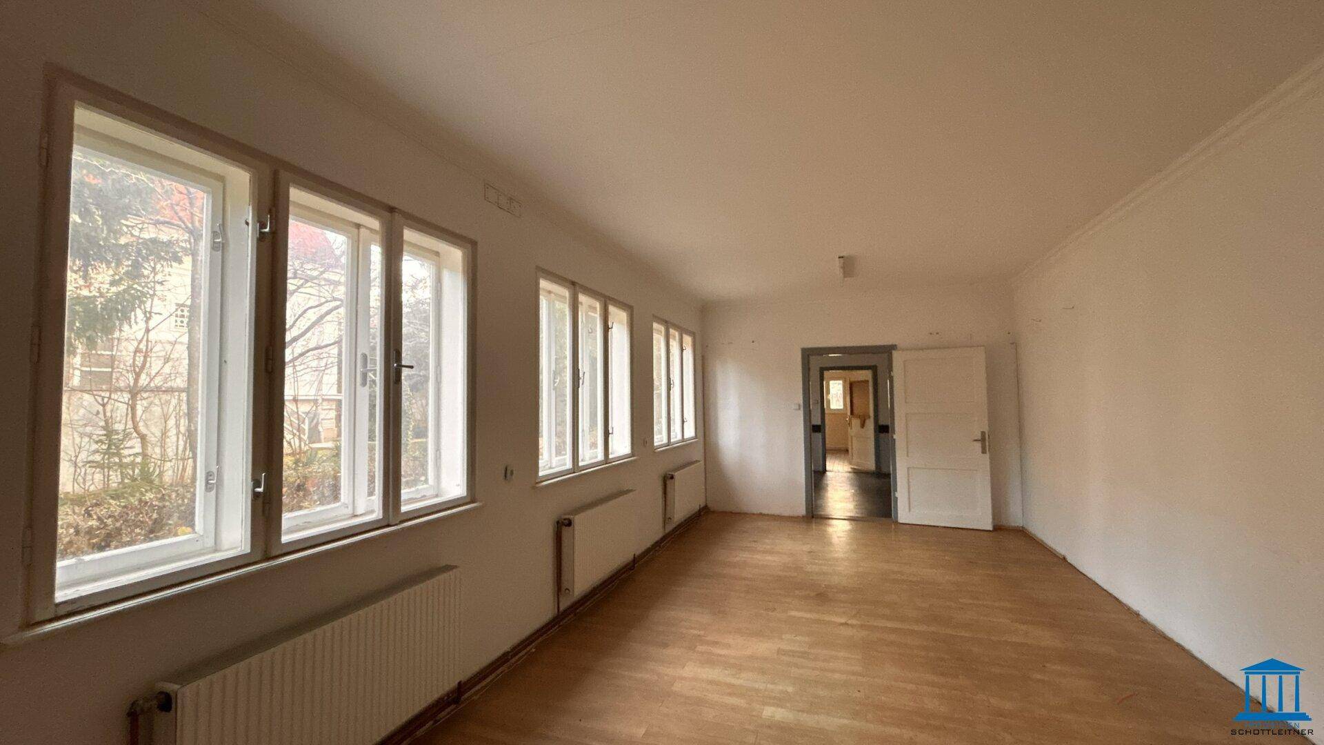 Heller, leerer Raum mit Holzfußboden und einer Reihe von Fenstern, die den Blick nach draußen freigeben.