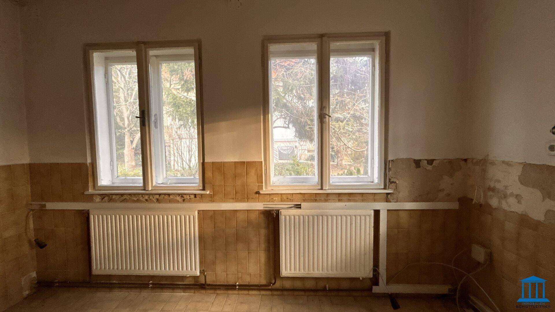 Raum mit teilweise gefliesten Wänden und zwei Fenstern, der eine umfassende Renovierung benötigt.