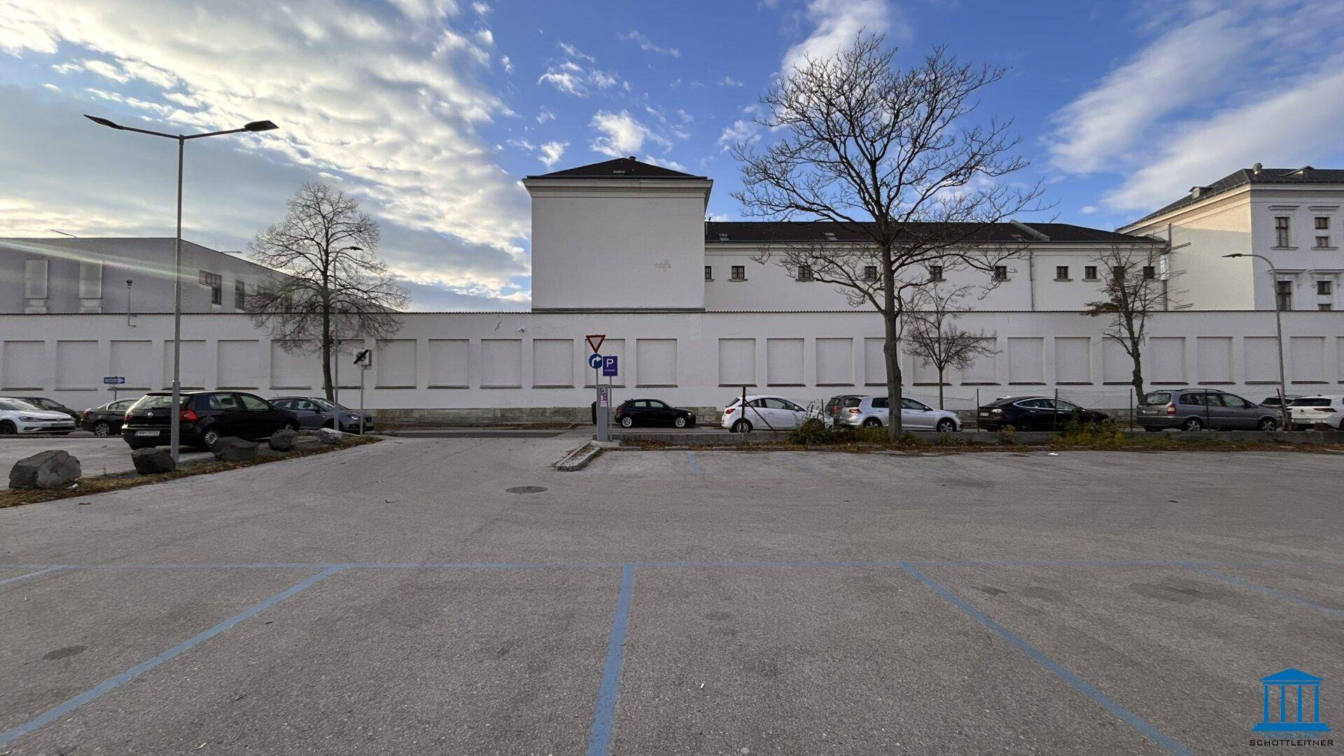 Parkplatz mit blauen Markierungen vor einer langen weißen Mauer und Gebäuden im Hintergrund.