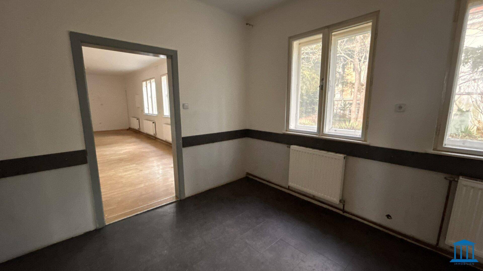 Leerer Raum mit dunklem Boden und Blick durch eine offene Tür in einen weiteren Raum.