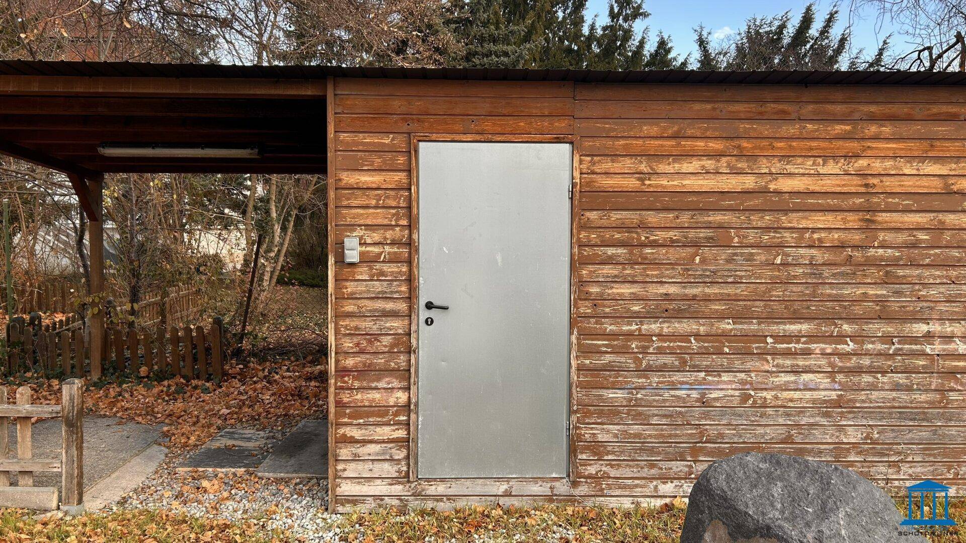 Holzschuppen mit einer einfachen Metalltür und einem angrenzenden überdachten Bereich.