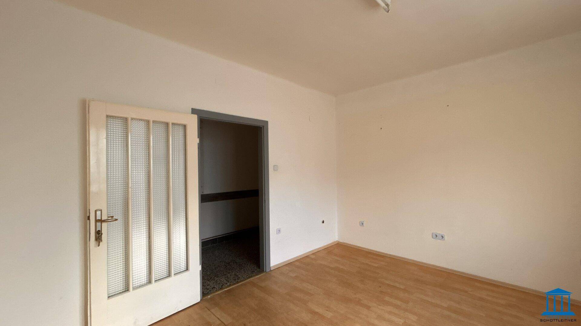 Leerer Raum mit Holzfußboden und einer Tür mit Glaseinsätzen, die in einen weiteren Raum führt.