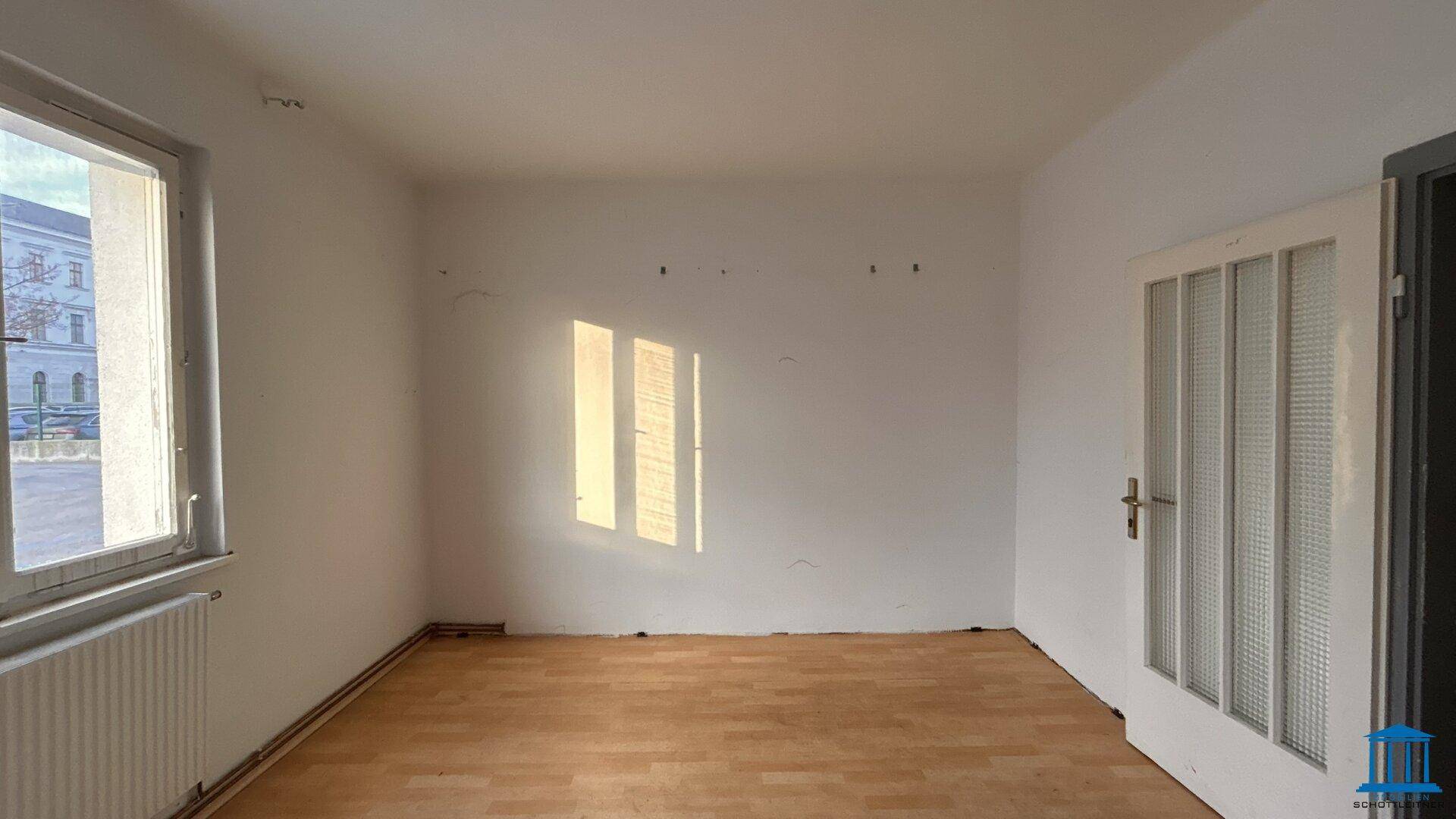 Leerer Raum mit Holzfußboden und einem Fenster, das Sonnenlicht auf die weiße Wand wirft.