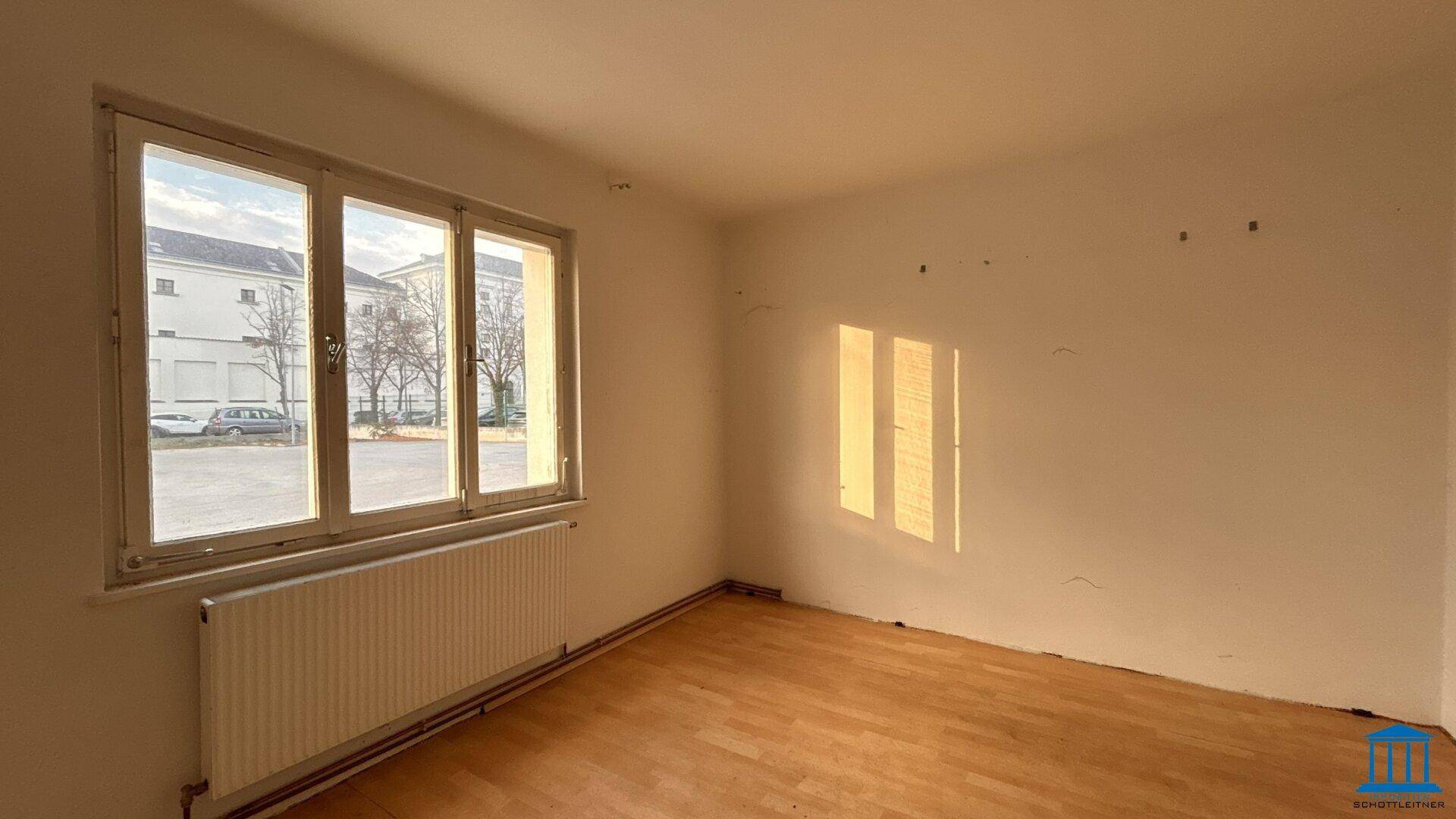 Heller, leerer Raum mit Holzfußboden und einem großen Fenster, das viel Tageslicht hereinlässt.