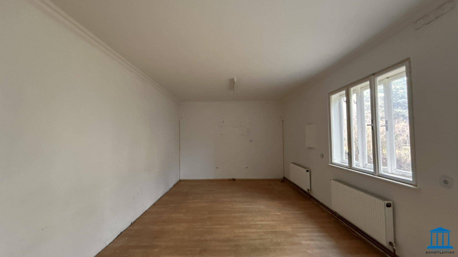 Leerer Raum mit Holzfußboden, weißen Wänden und einem Fensterbereich mit Heizkörpern.